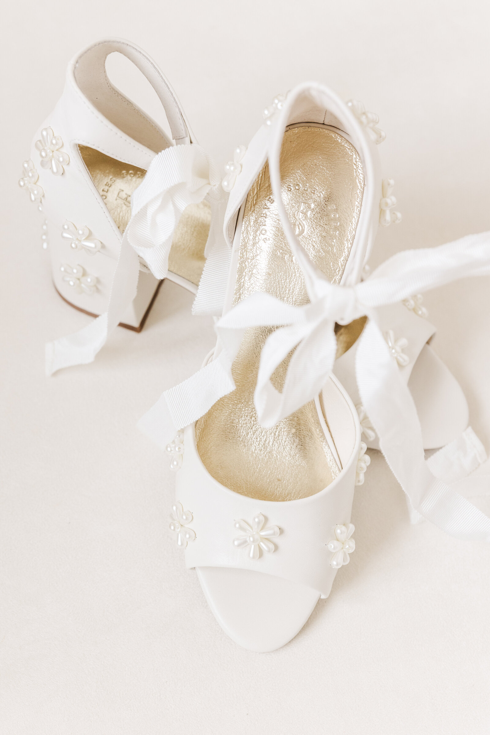 Flat lay of white bridal block heels adorned with floral pearl appliqués and tied with soft white ribbon bows, styled on a light neutral background for an elegant wedding day detail shot.