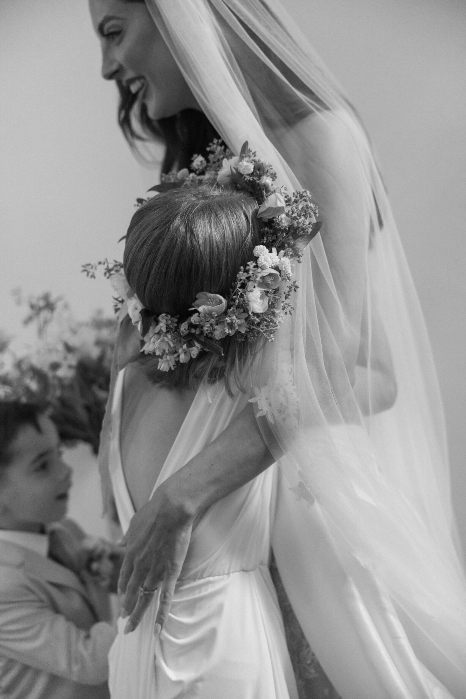 Black & white candid photo of bride with draping veil hugging young daughter in flower crown and younger son