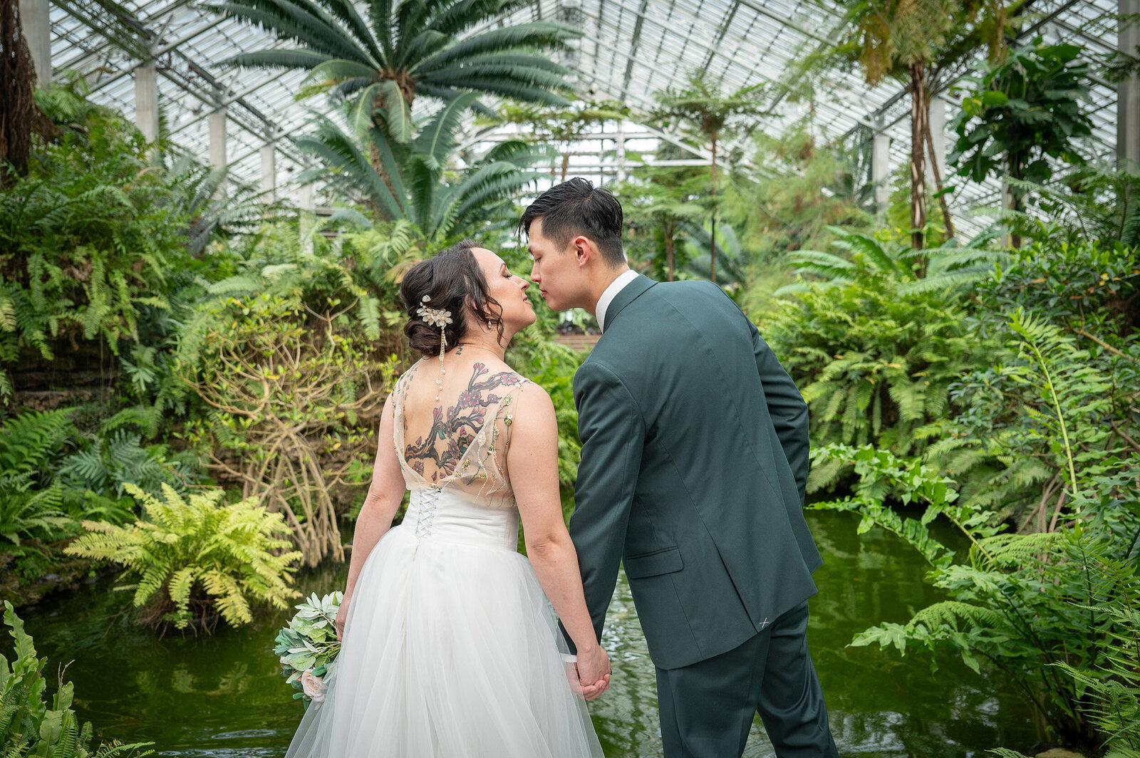 bride and groom leaning in to kiss surrounded by a greenhouse and near a pond