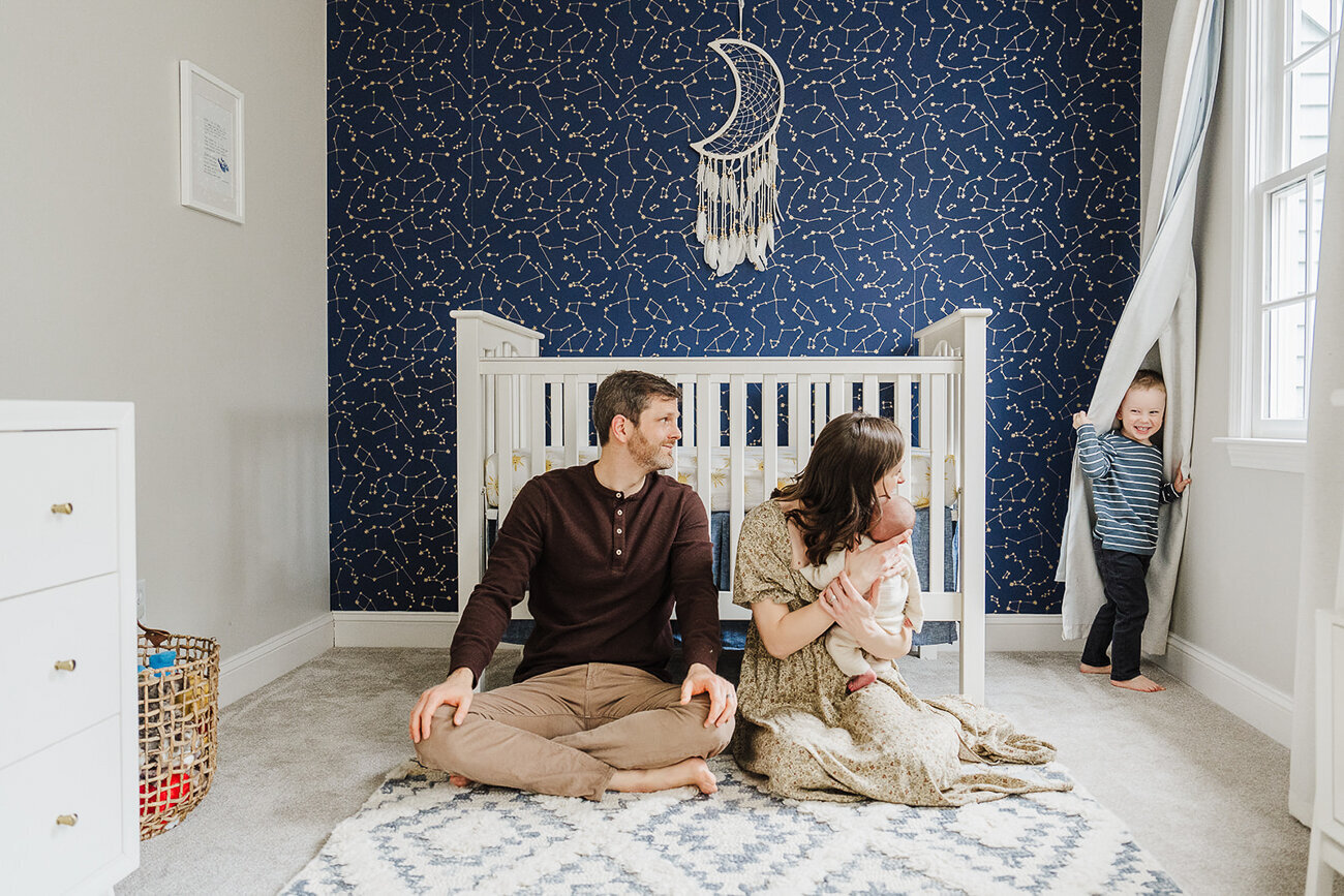 big brother hides behind nursery curtain while parents snuggle newborn