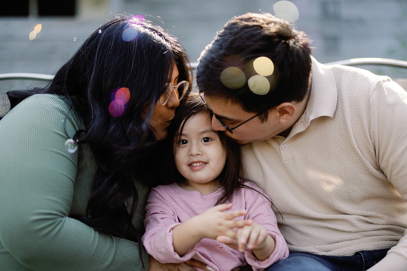 A soft-lit family portrait of parents kissing their young daughter on the cheek, framed by lens flares and a warm outdoor ambiance.