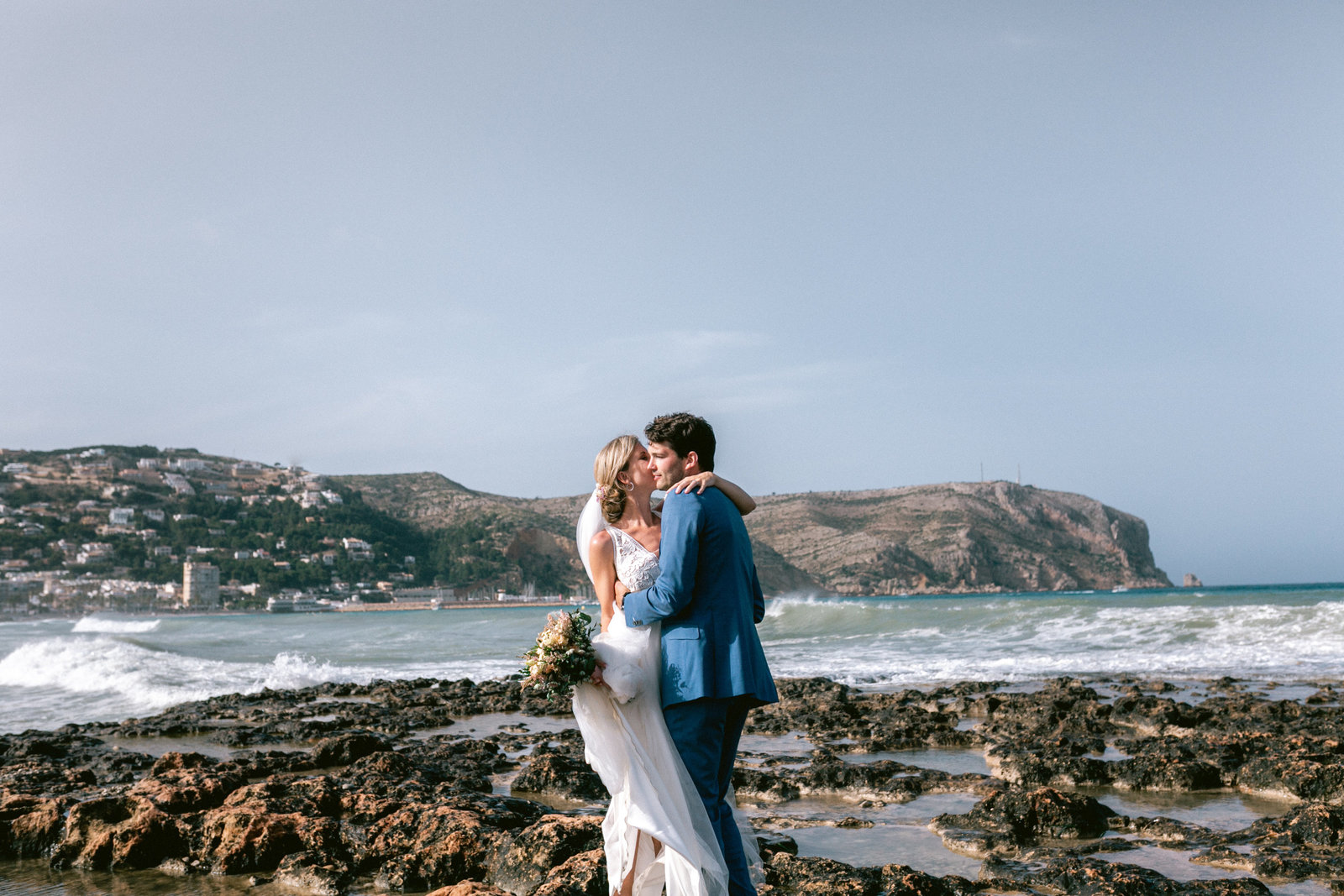 Bruidspaar dat elkaar omhelst op het strand tijdens hun bruiloft in la siësta Javea Spanje met landschap op de achtergrond
