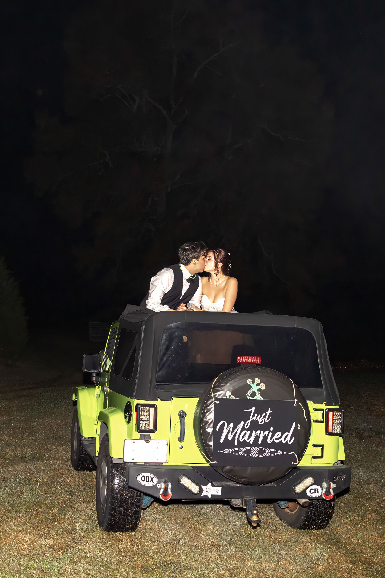 The bride and groom share a kiss in the back of a green "Just Married" Jeep at night, captured during a Wilmington, NC wedding, showcasing fun and adventurous wedding day details.
