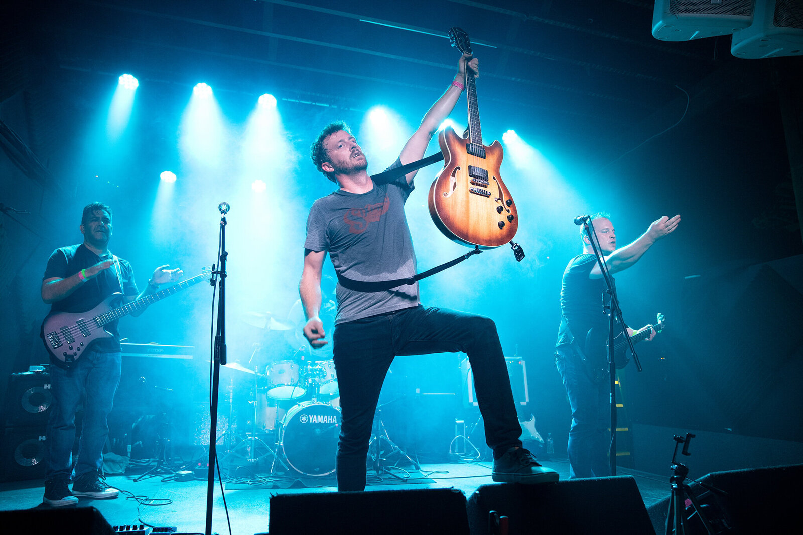 A rock band performs energetically on stage, blue lights shining behind them. Captured by a skilled Jacksonville band photographer, the lead guitarist raises his guitar high while others play guitar, bass, and drums in this dynamic concert photography Jacksonville moment.