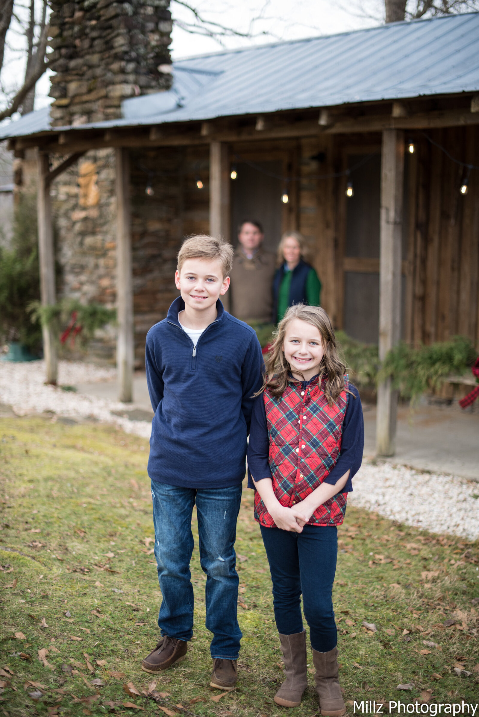 a brother and sister standing in front of a house smiling at the camera while their parents watch from the background photographed by Millz Photography in Greenville, SC
