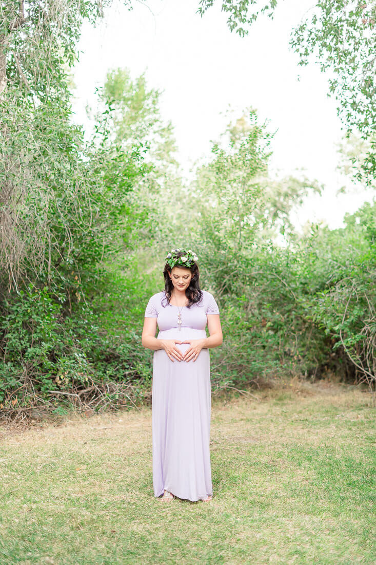 A pregnant woman wearing a long lavender maternity dress and a flower crown looks down at her belly as she stands in a small grove of trees at Highland Glenn Park in the summer