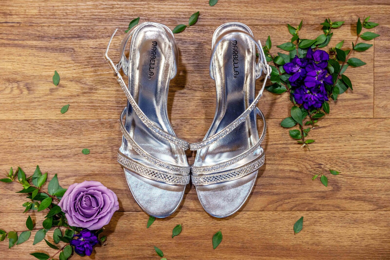 A pair of sparkling silver bridal heels surrounded by purple roses and greenery, captured on wooden flooring during a Boone, NC wedding, blending elegance and personalized bridal details.