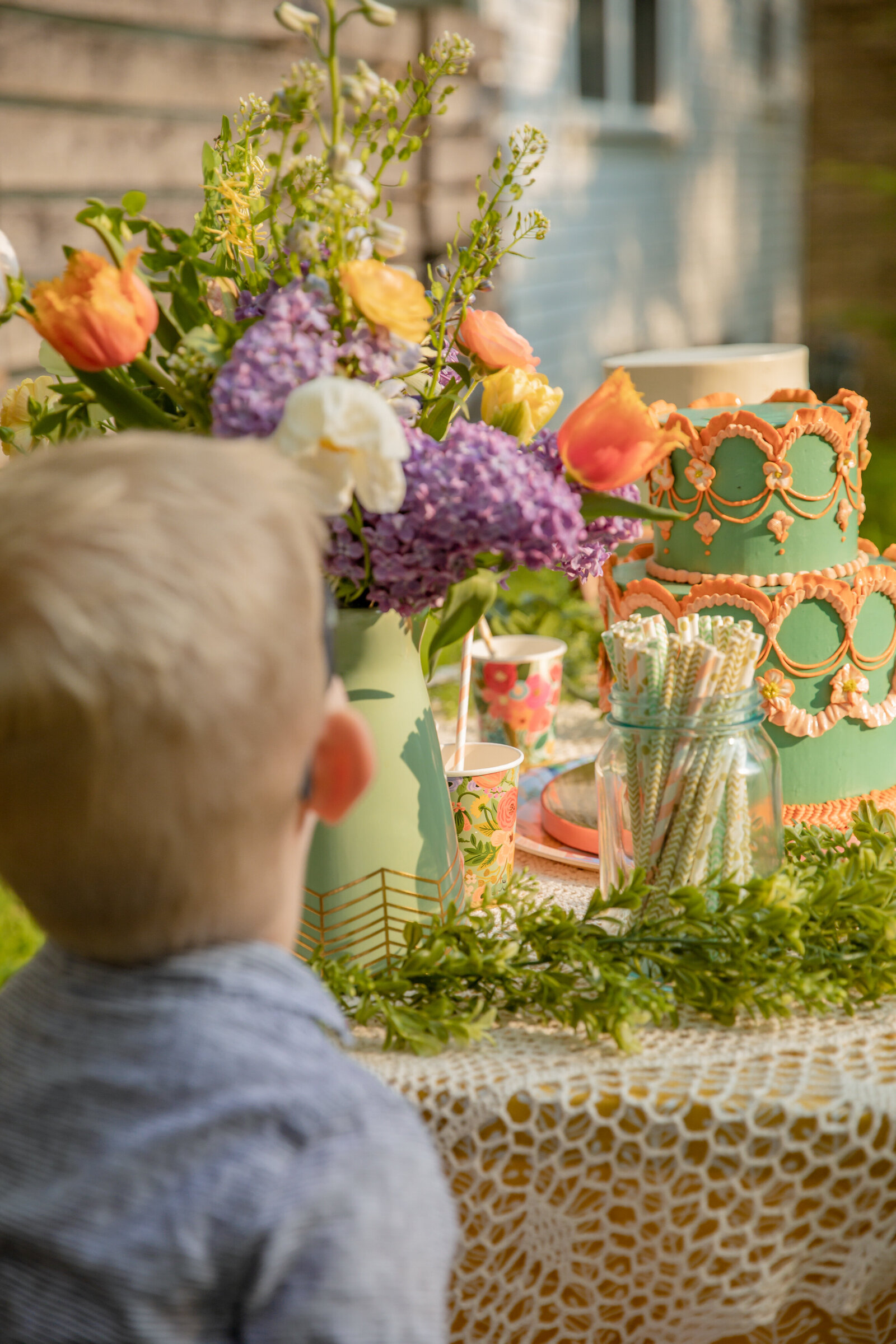 toddler boy looks at table full of cake and flowers for garden party styled session, shot from behind over shoulder