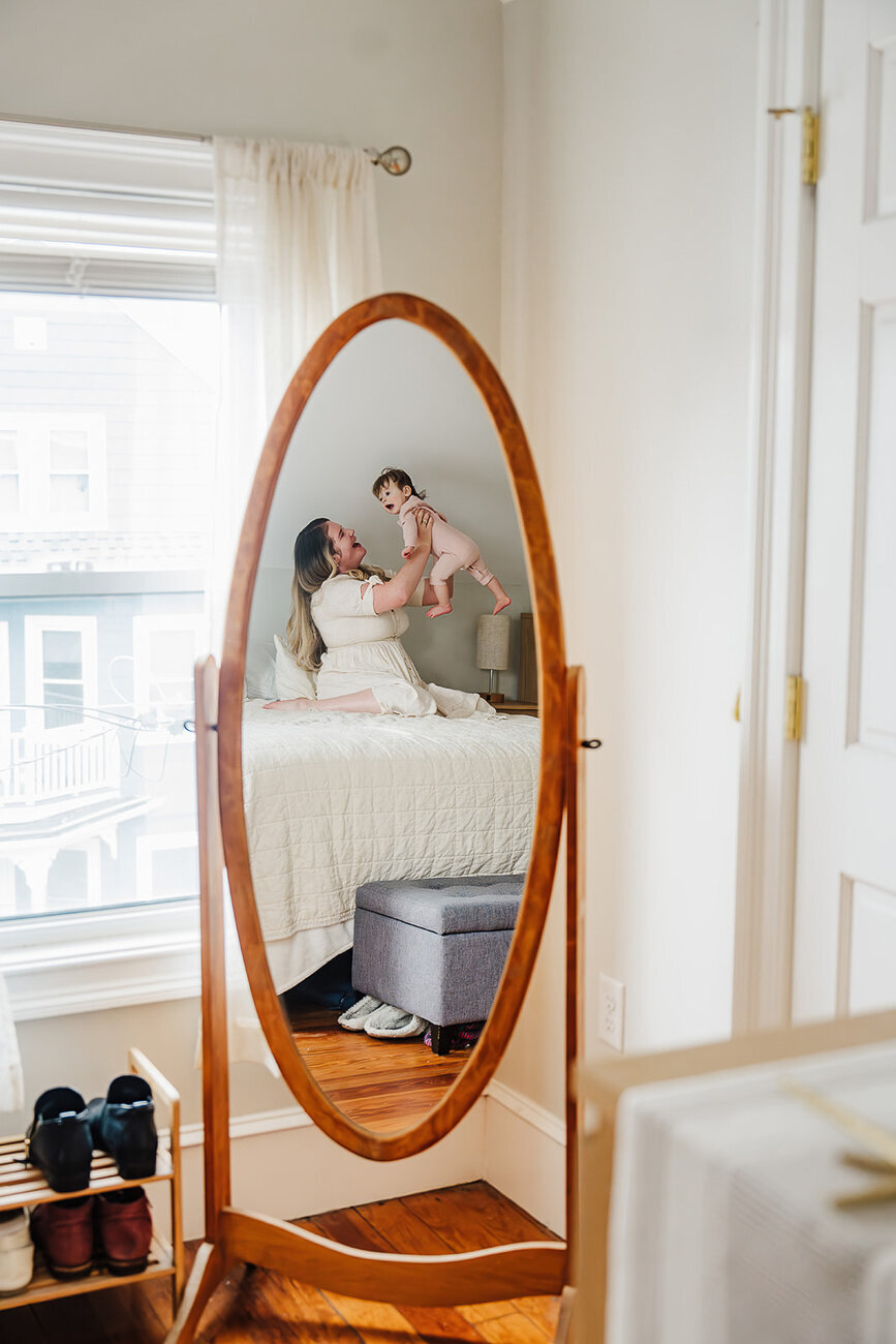 reflection in a mirror of a mom holding toddler in air on bed in home