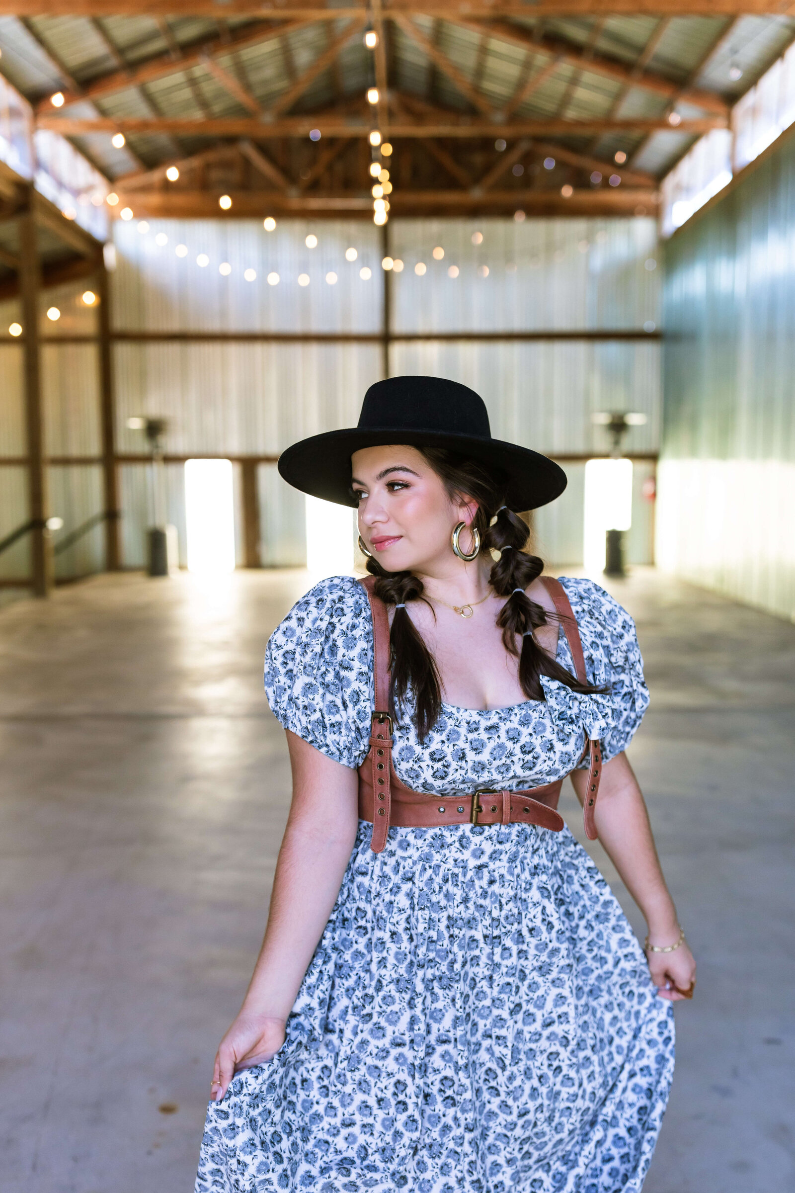 dark haired girl wearing black hat, brown belt, and blue floral dress with bubble braids in a warehouse