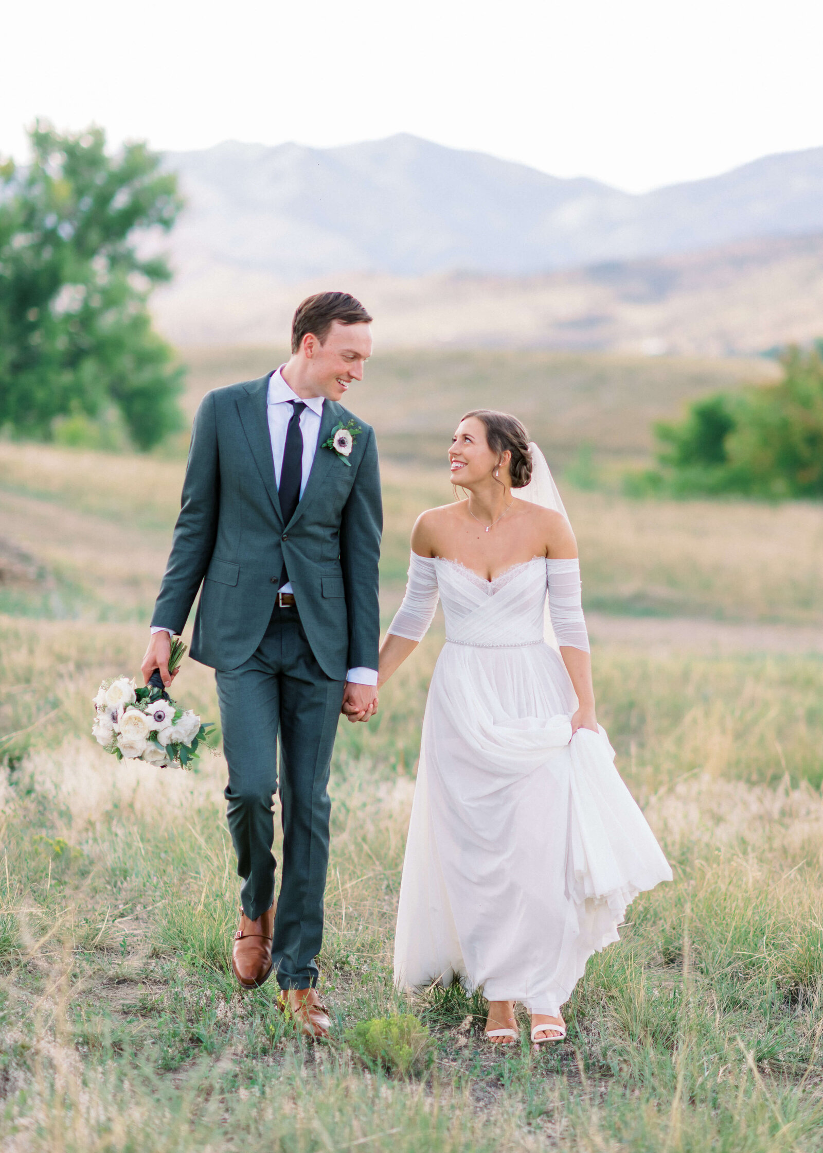 groom in green suit and bride in dress walking through a field with the mountains in the background