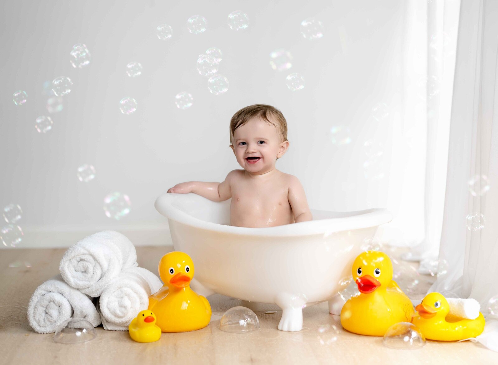 Baby boy smiling in white tub surround by rubber ducks and bubbles. Photographed at Atlanta baby photography studio.