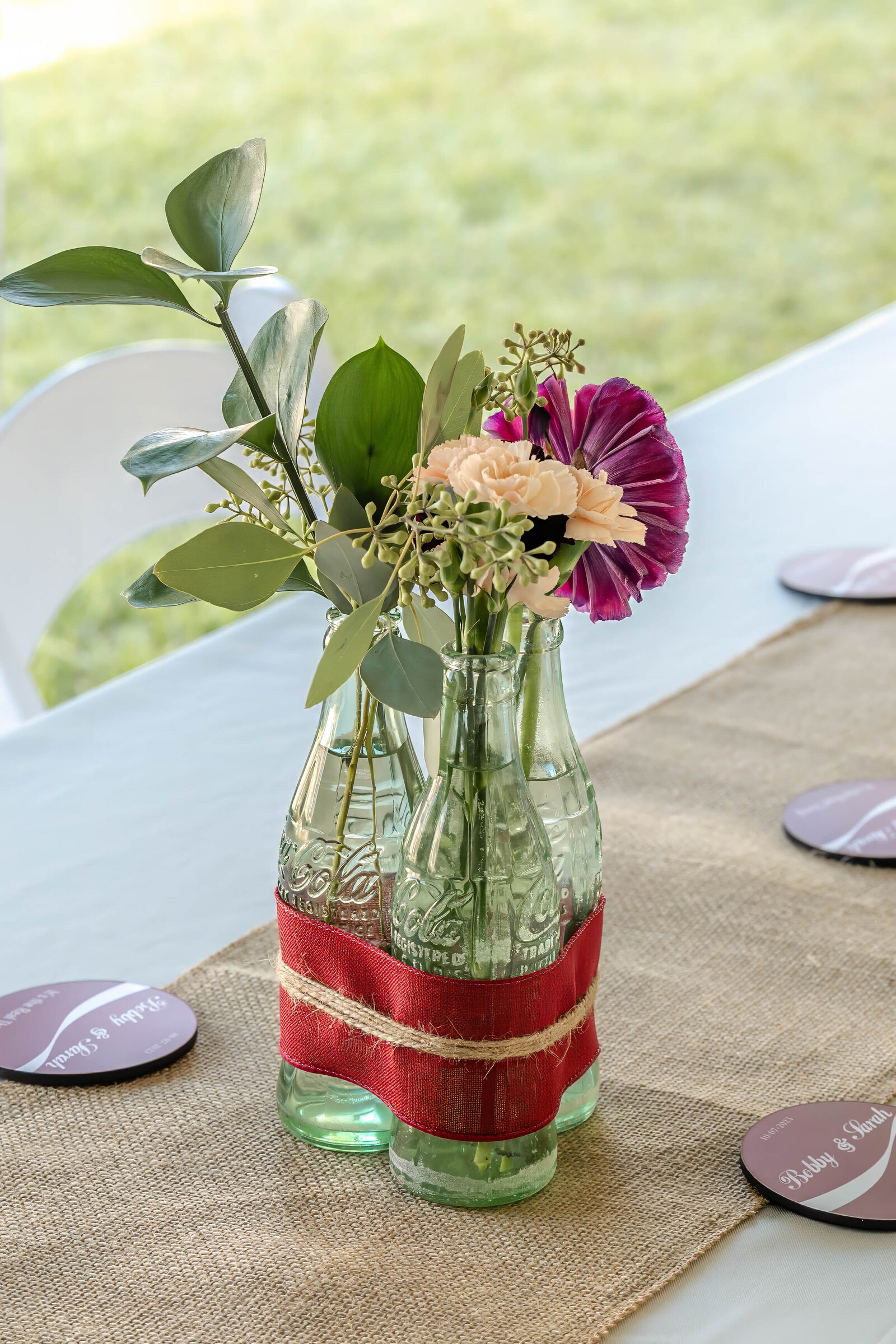 A rustic wedding centerpiece featuring green glass bottles wrapped in red fabric and filled with greenery and pink flowers, displayed on a burlap table runner, captured during a Wilmington, NC wedding.