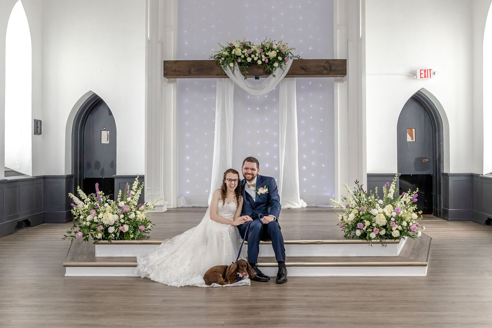 The bride and groom pose with their dog in front of a beautifully decorated altar with floral arrangements, captured during an elegant wedding in Asheville, NC, blending high-end style with authentic moments.