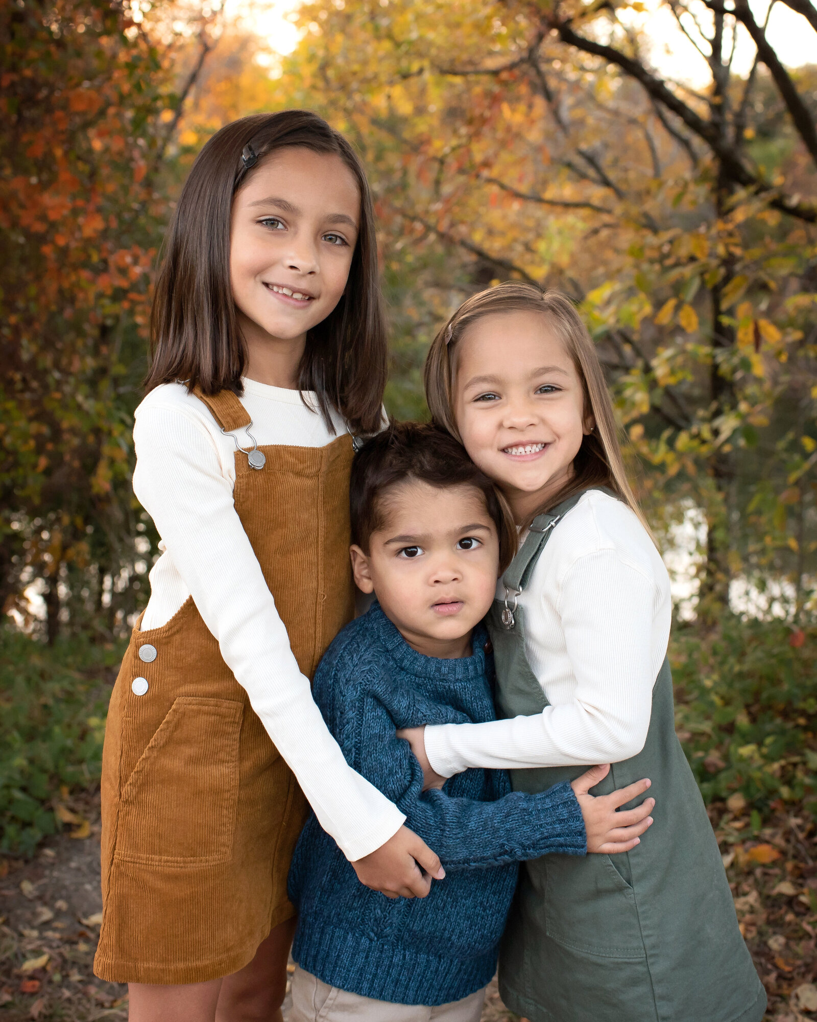 Three siblings with their arms around each other in front of colorful trees in Allen, TX.