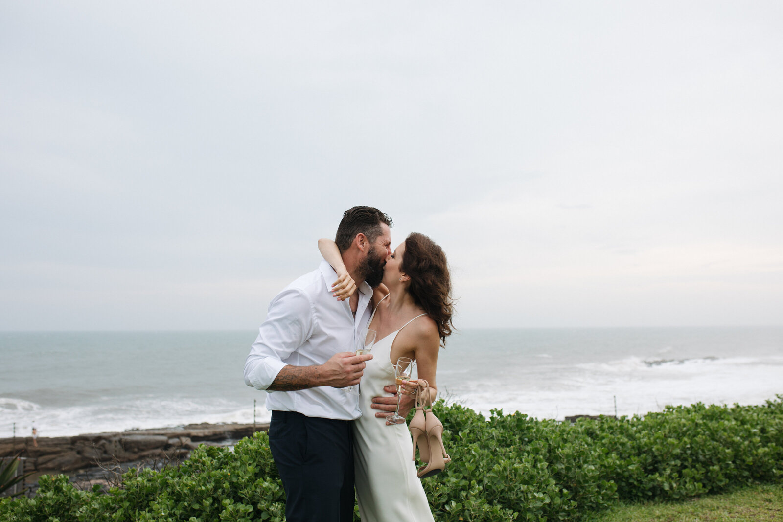 A bride and groom embrace while holding champagne , the ocean is in the background