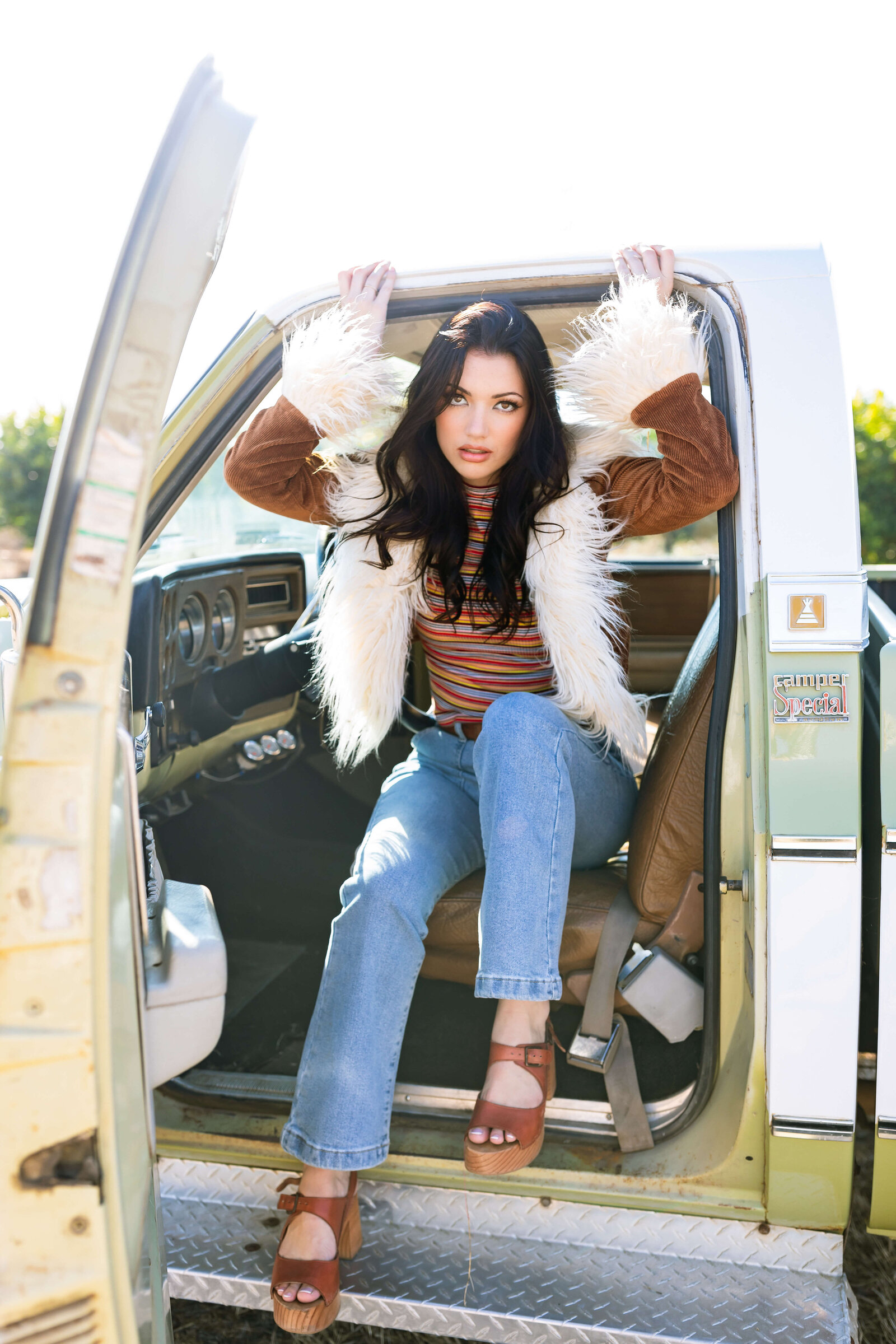 dair haired girl wearing brown fur coat, striped shirt, brown heels and posing while sitting in a green truck