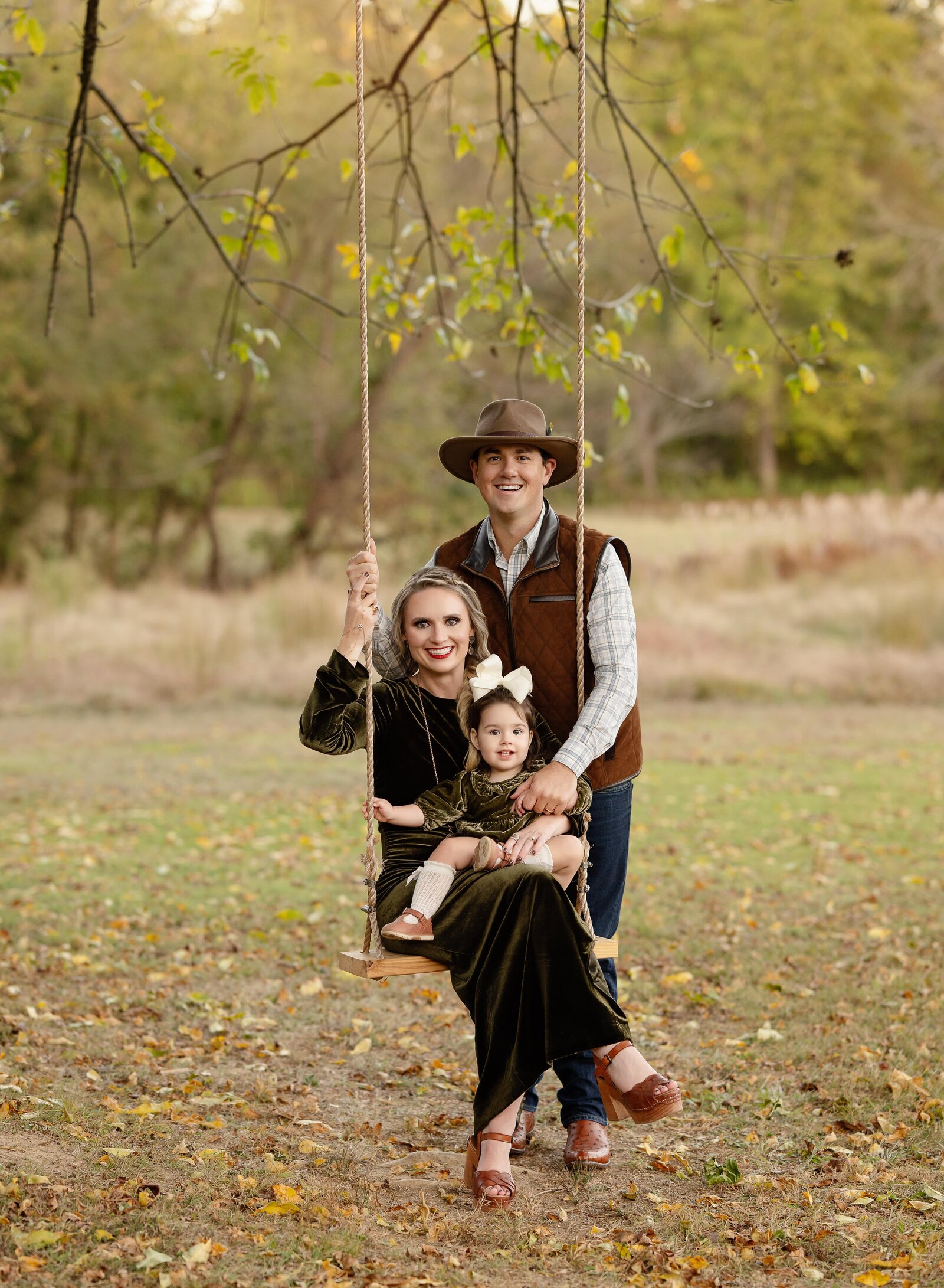 Family of 3 taking fall photos and sitting on a tree swing, smiling.