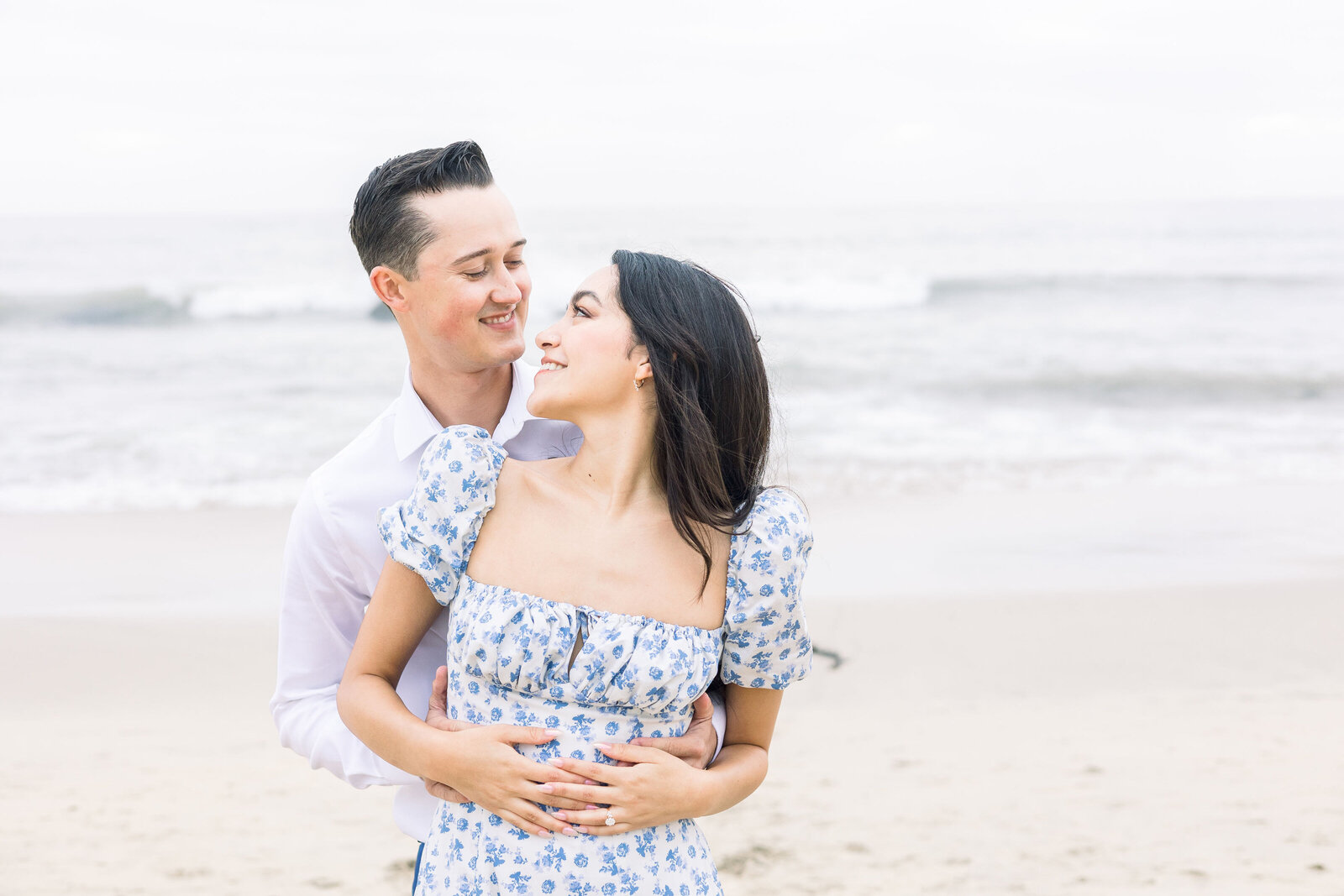 couples photos captured by wedding photographer bay area at half moon bay for beach engagement photos with the shore behind them as the man holds the womans wait while she looks back to him