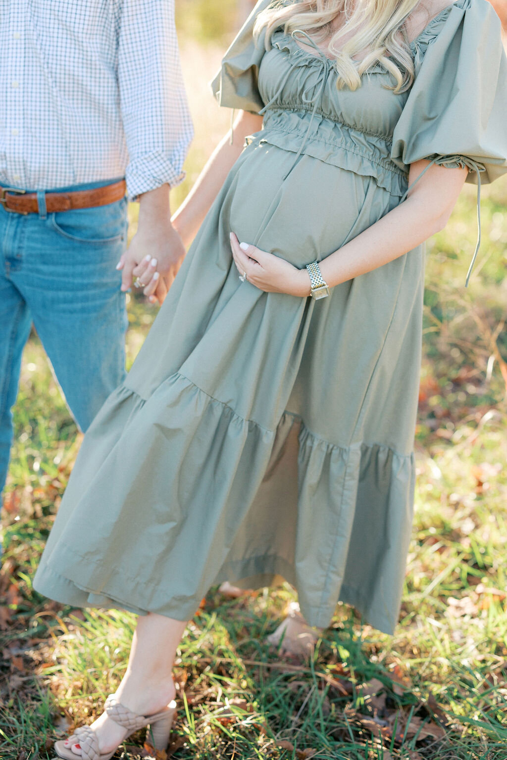Pregnant woman in green dress holds baby bump walking through field with husband