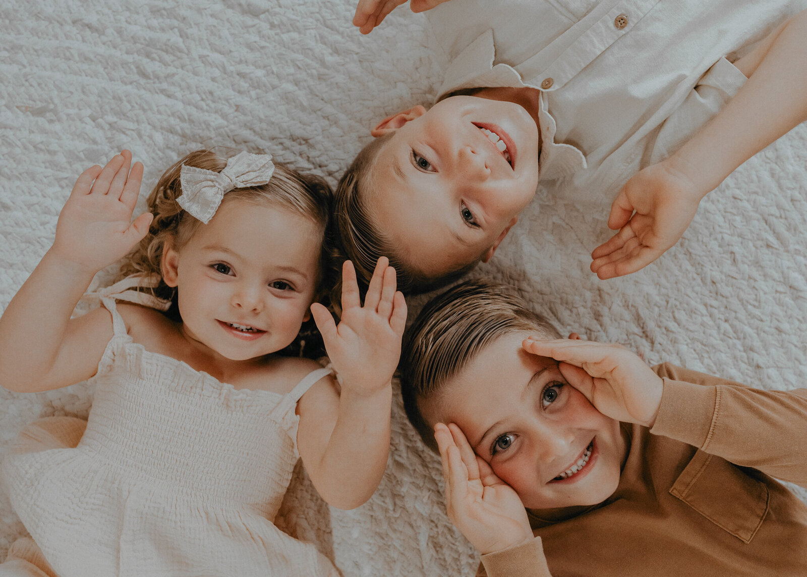 Three young siblings, make goofy faces at the audience while laying on the floor with their heads together.