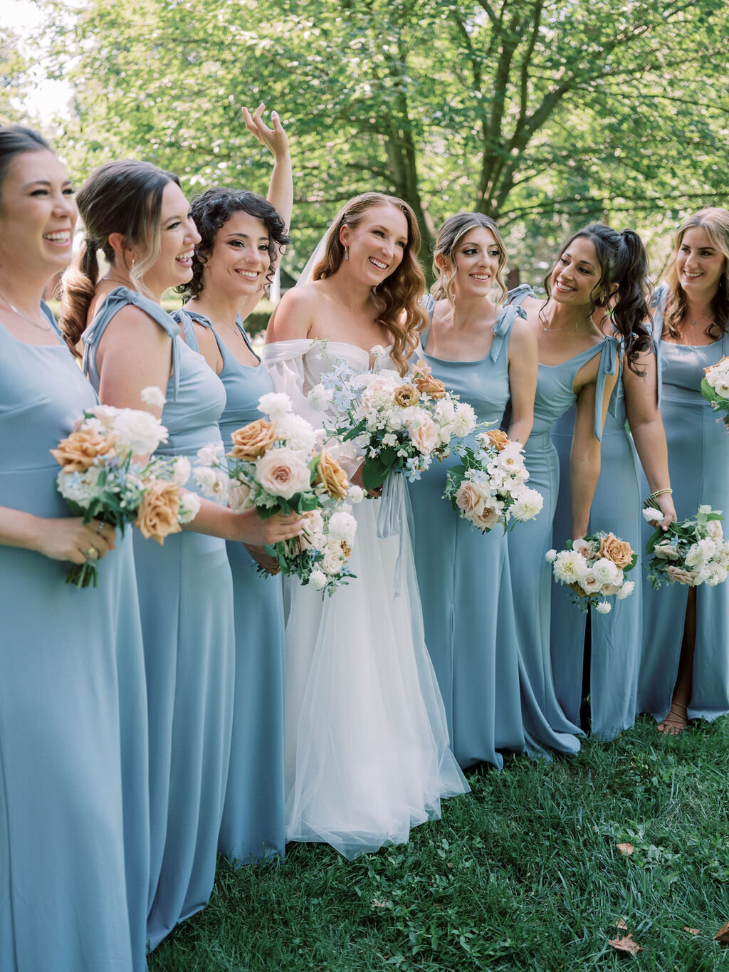 Bride and bridesmaids in blue dresses holding bouquets of peach garden rose, white dahlia, toffee roses, white hydrangea, light blue delphinium and brown lisianthus.