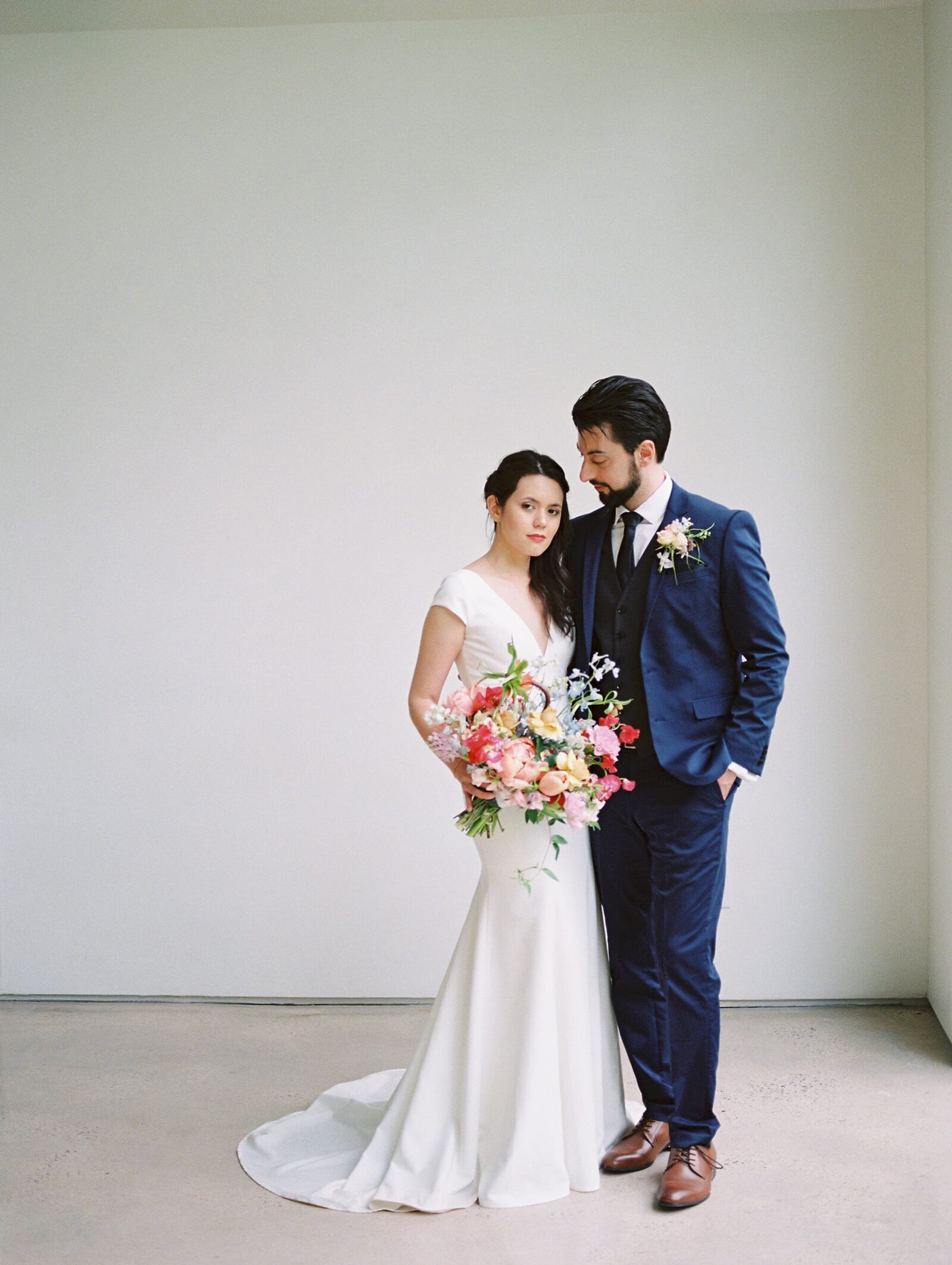 A bride and groom stands close together, the bride holding a large, colorful, Dutch Masters inspired bouquet with a clean white background.