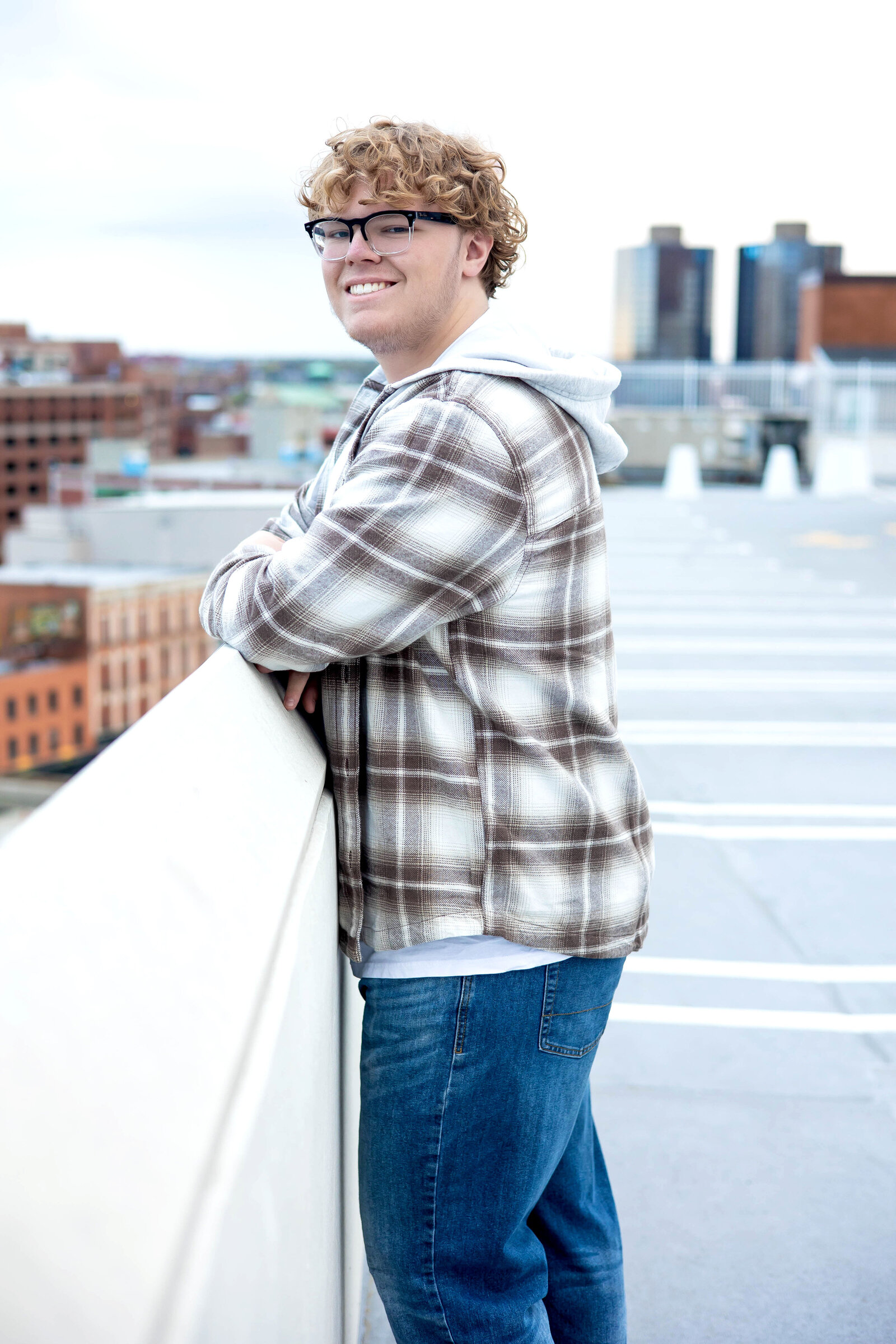 Caucasian senior boy wearing a plaid fleece jacket, white shirt, and blue jeans leaning against a ledge on the rooftop of a parking garage.