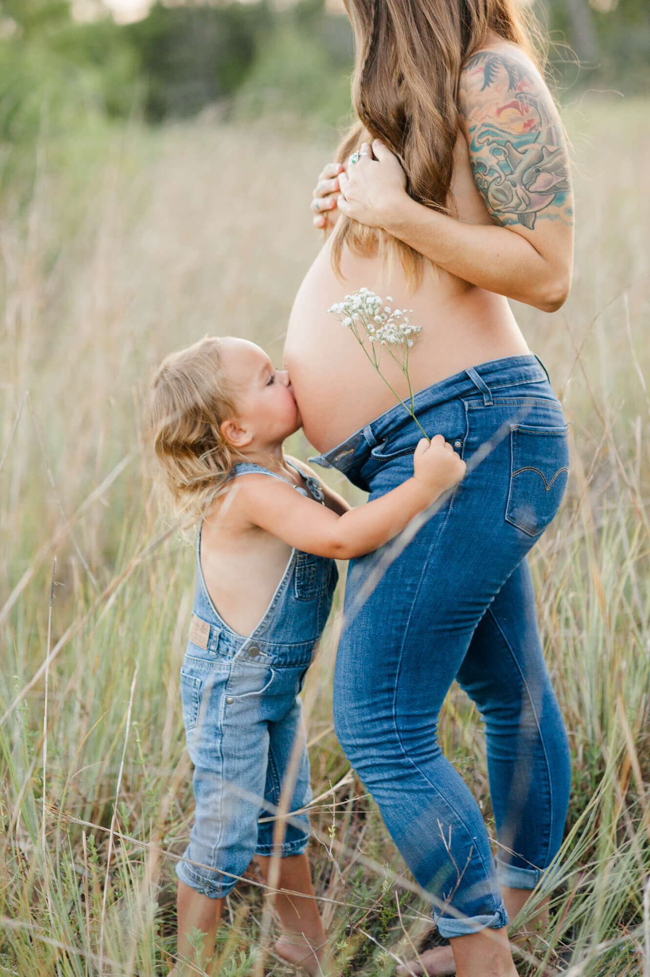 Pregnant mom stands topless in a tall grass field covering her breasts while her young daughter kisses her pregnant belly in jeans