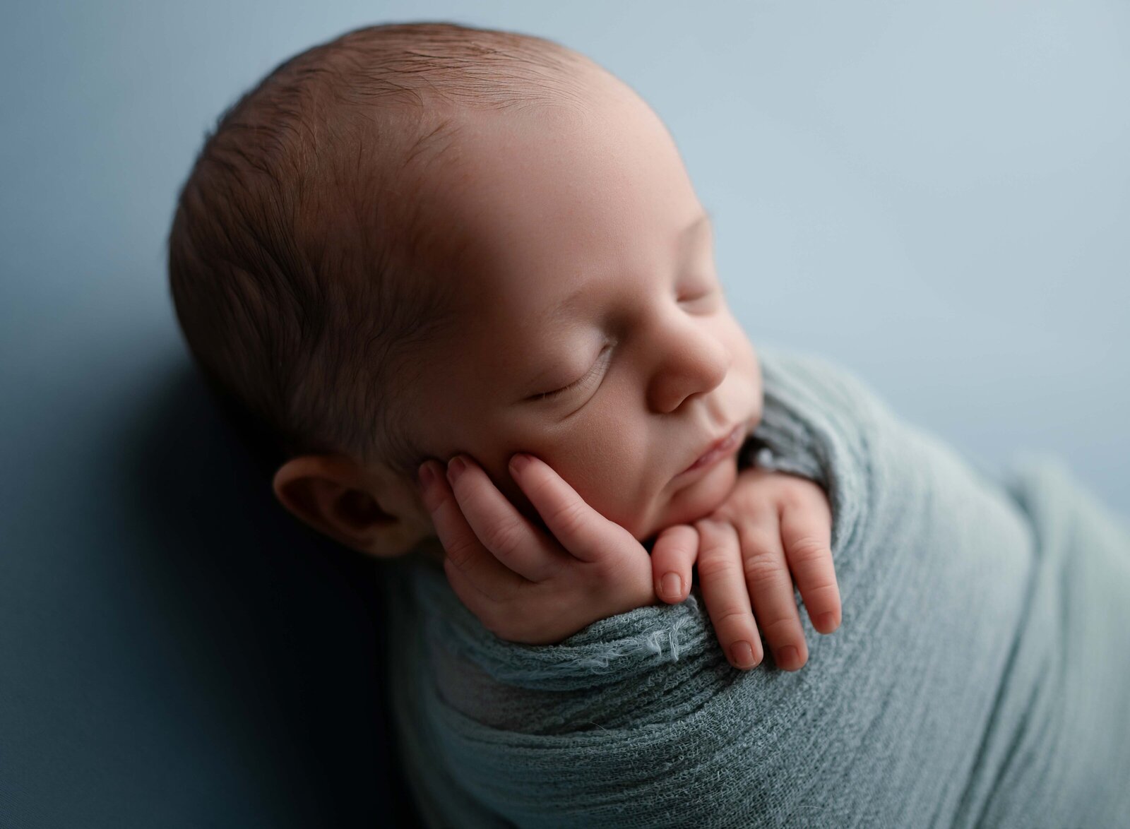 Baby boy swaddled in blue with hand on cheek, sleeping. Photo taken by Jenni Nikki at Newborn photo studio in Marietta, GA.