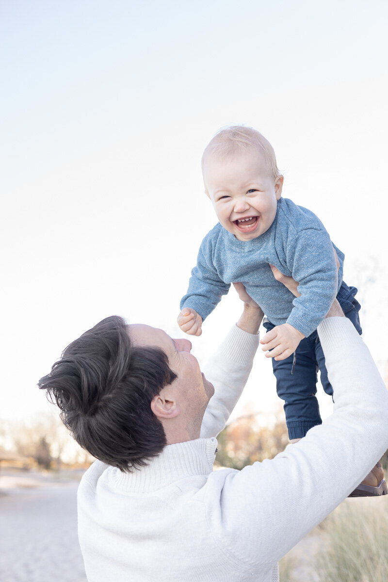 Smiling baby boy being lifted in the air by his dad during a candid family photo session in Fairfield County, CT. Captured by Nicole, a Westchester and Connecticut lifestyle family photographer known for documenting joyful and natural family moments.
