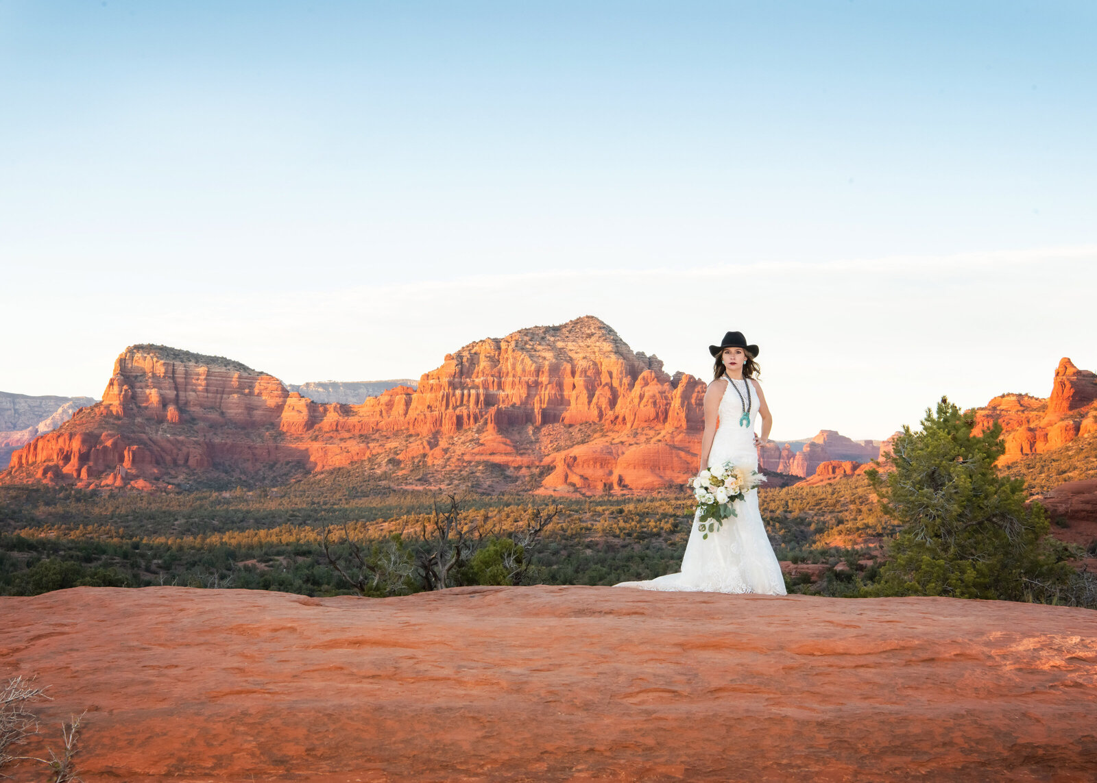 Fearless destination bride standing in the Sedona dessert in her white wedding dress holding a bouquet and wearing a cowboy hat at Bell Rock