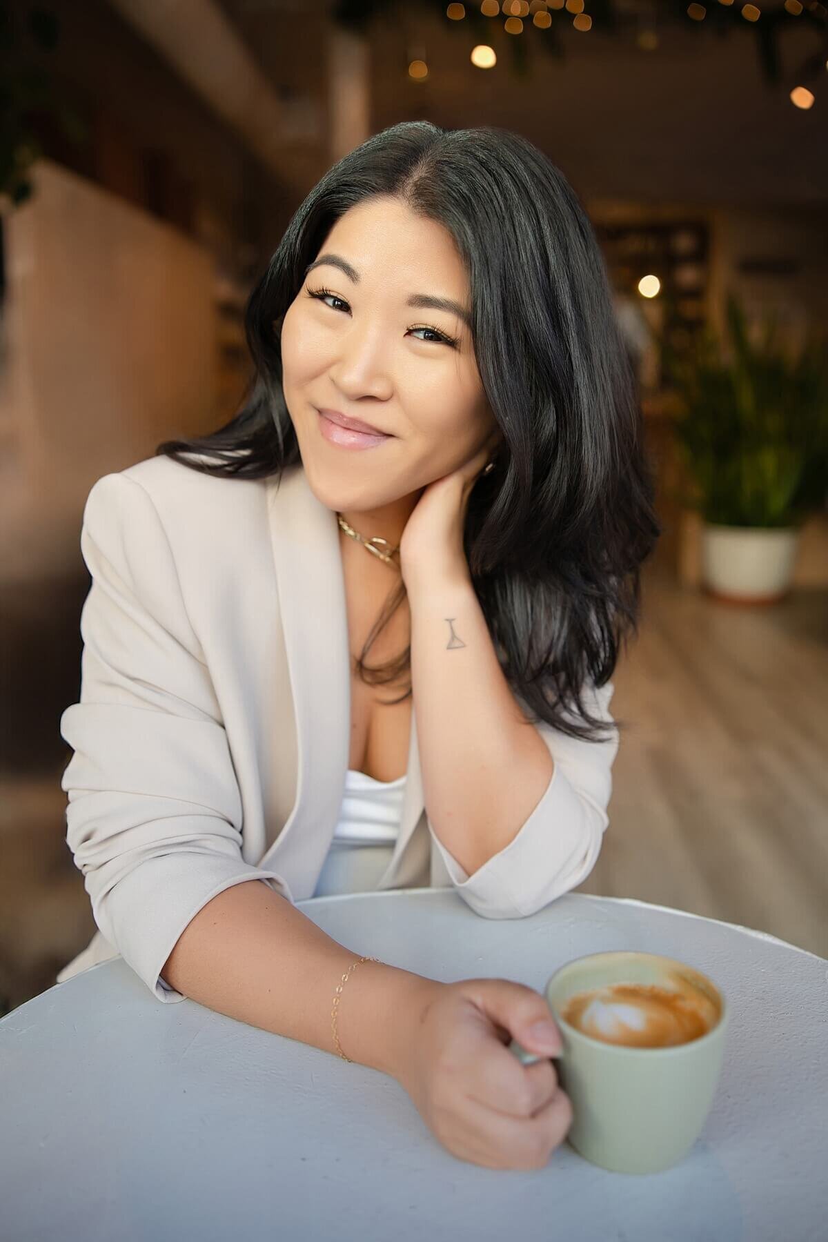 Personal branding photo of an Asian woman entrepreneur smiling confidently at a café table with a latte, wearing a light blazer and gold necklace. Captured by a Seattle brand photographer, this image conveys warmth, professionalism, and approachability for business owners building personal brands.