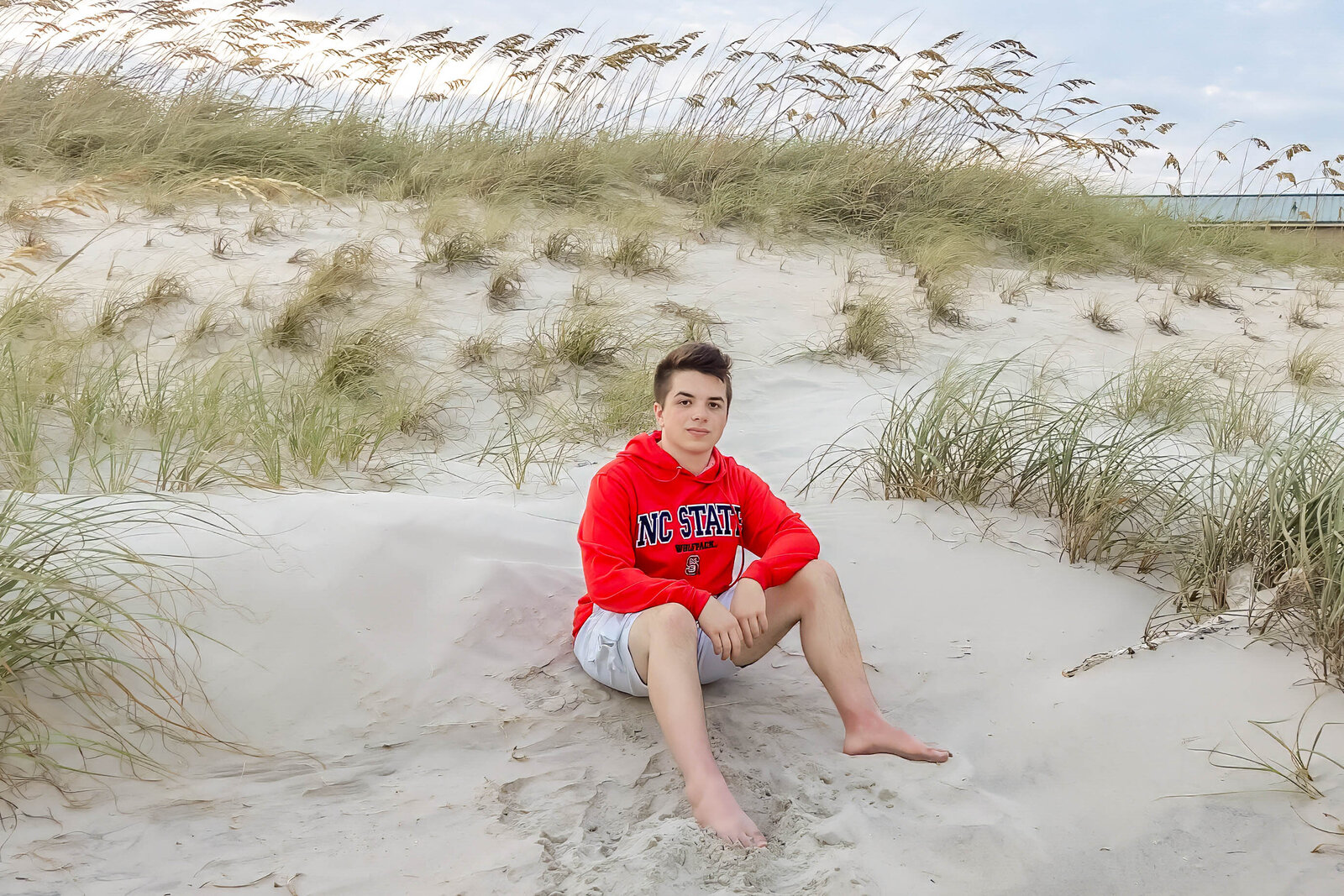 A high school senior in a red hoodie sitting on sand dunes with sea oats in the background in Topsail Beach, North Carolina. This relaxed portrait highlights the natural beauty of outdoor senior photography, perfect for beach-themed sessions.