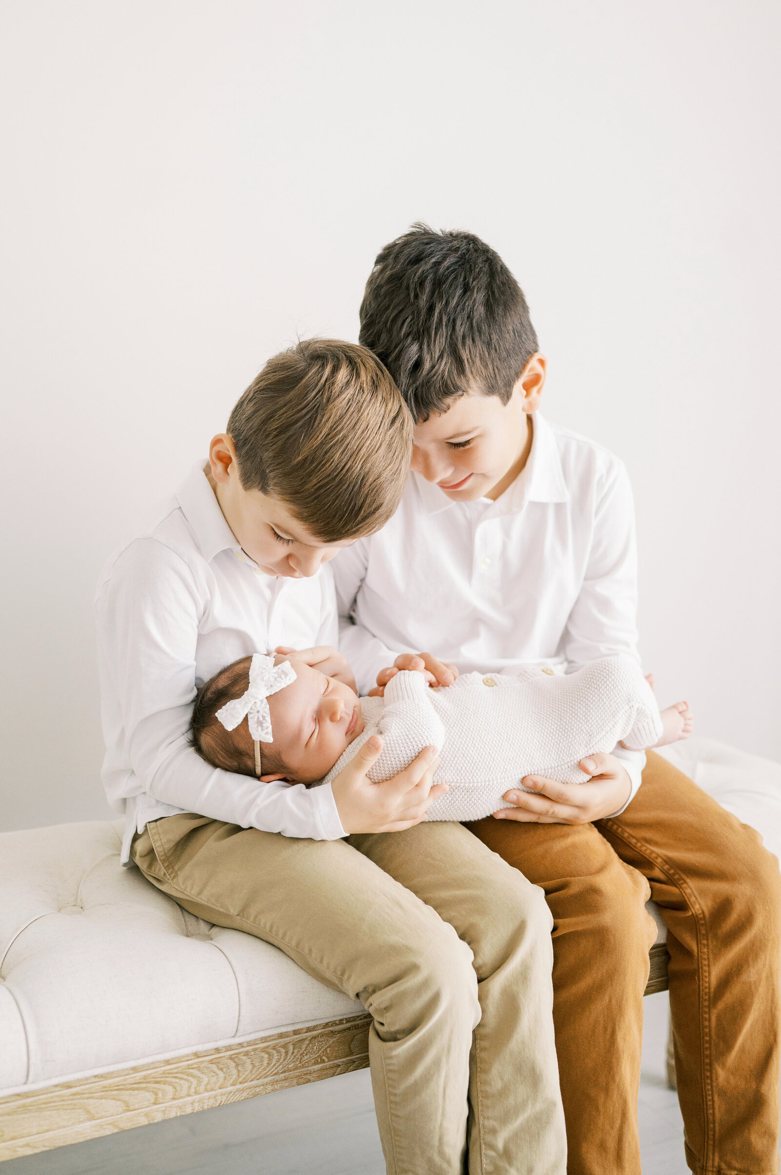 Two brothers in white shirts sit on bench holding newborn baby sister during photography session in Raleigh NC