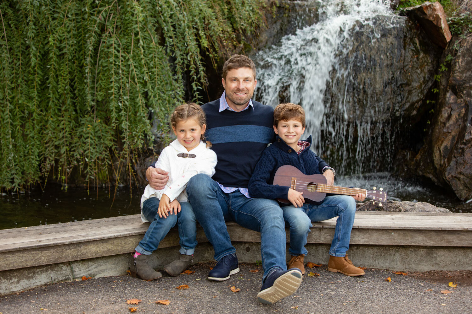 A man is sitting on a bench with two children and holding a guitar in front of a waterfall.