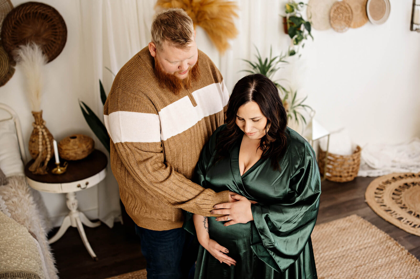 A man stands beside a pregnant woman in a green dress, gently resting his hand on her belly. Captured by a Bloomington maternity photographer, the cozy indoor setting features plants, baskets, and woven rugs for timeless maternity portraits.