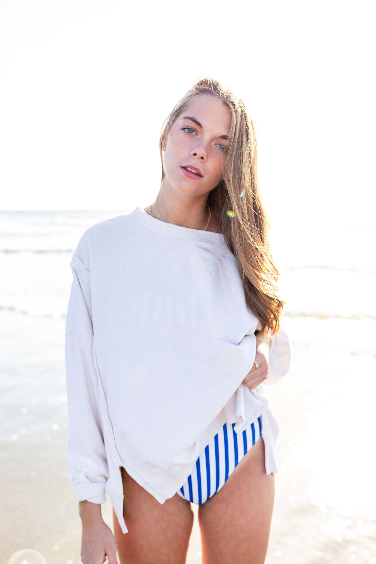 brunette high schooler posing on a beach wearing a white shirt and striped swimsuit