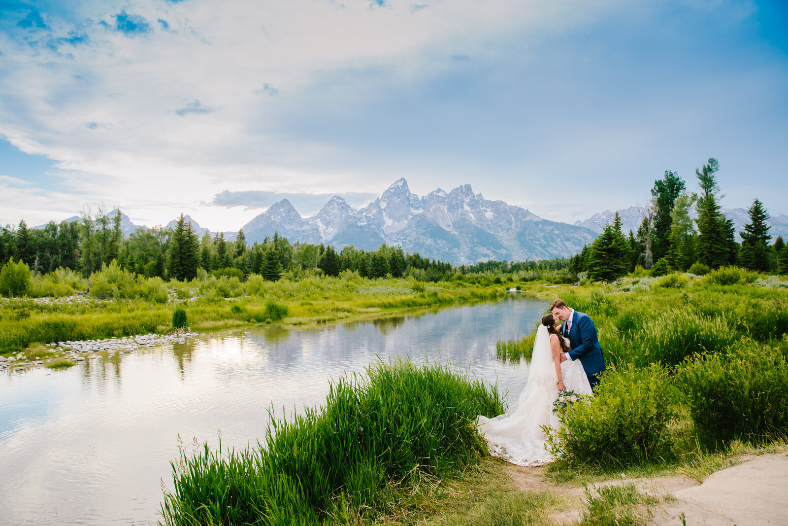 spring time jackson hole wedding at schwbachers landing in grand teton national park. The  bride and groom are kissing by the waters edge and the sky is bright blue