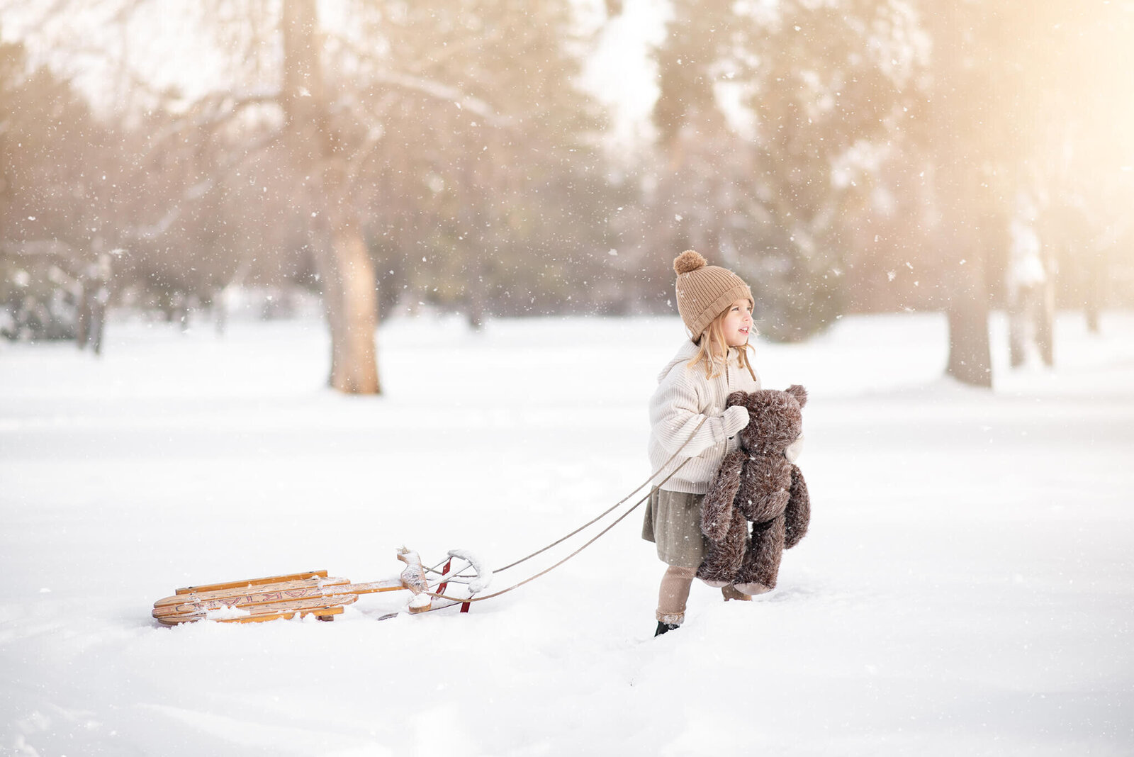 little girl holding a teddy bear in the snow, while pulling a wooden sled