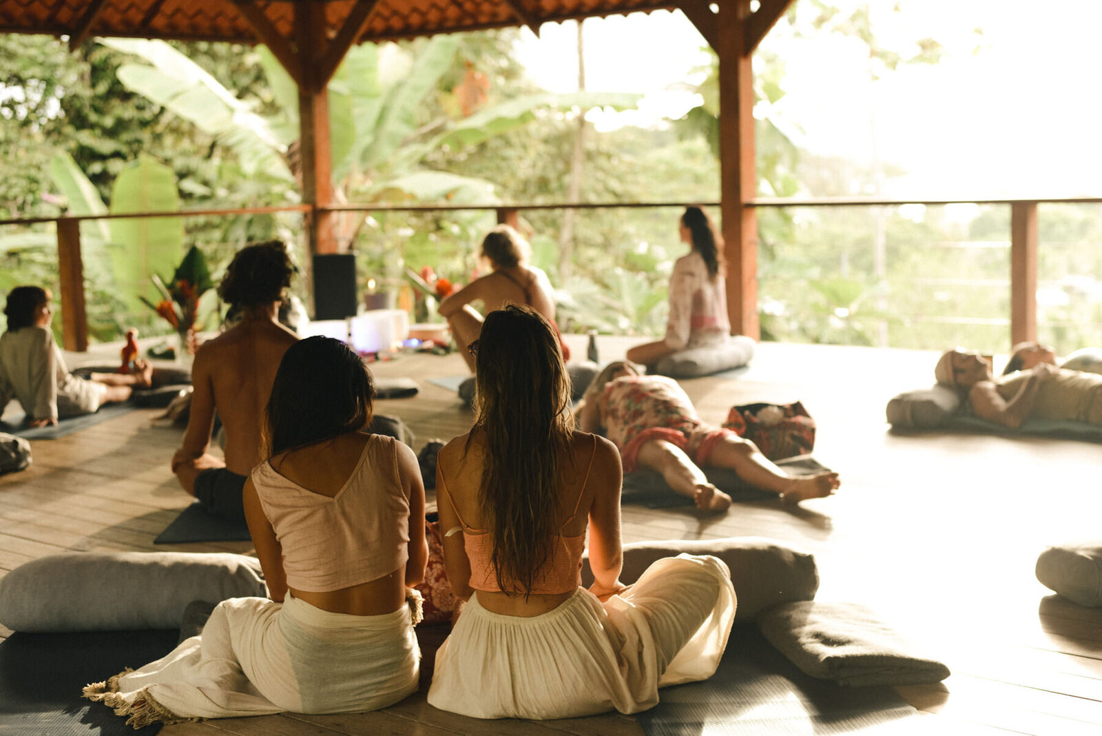 People sitting and lying on cushions in a wooden pavilion, surrounded by lush greenery. Sunlight filters through the open space, creating a serene atmosphere. Participants wear casual, comfortable clothing, engaged in a peaceful group activity.