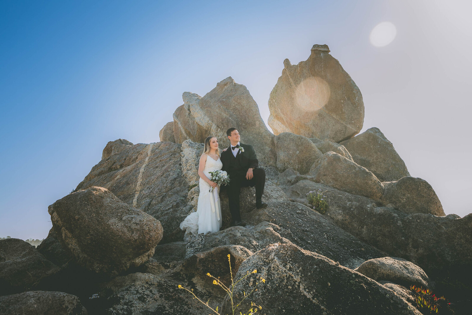 A bride and groom pose on rocks and look towards the right of the frame as a sun flare appears.