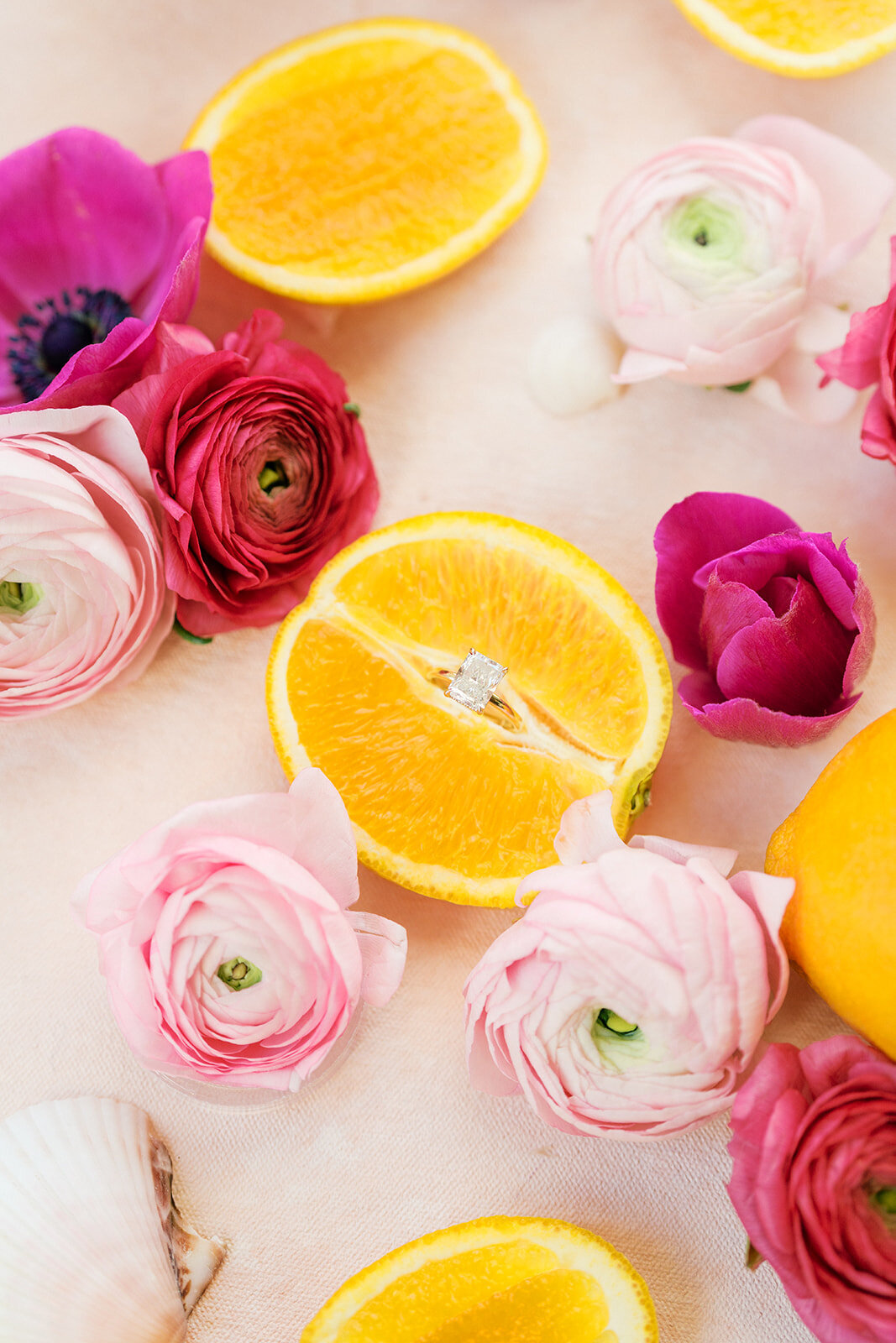 Bright pink and light pink flowers lay with sliced orange holding engagement ring