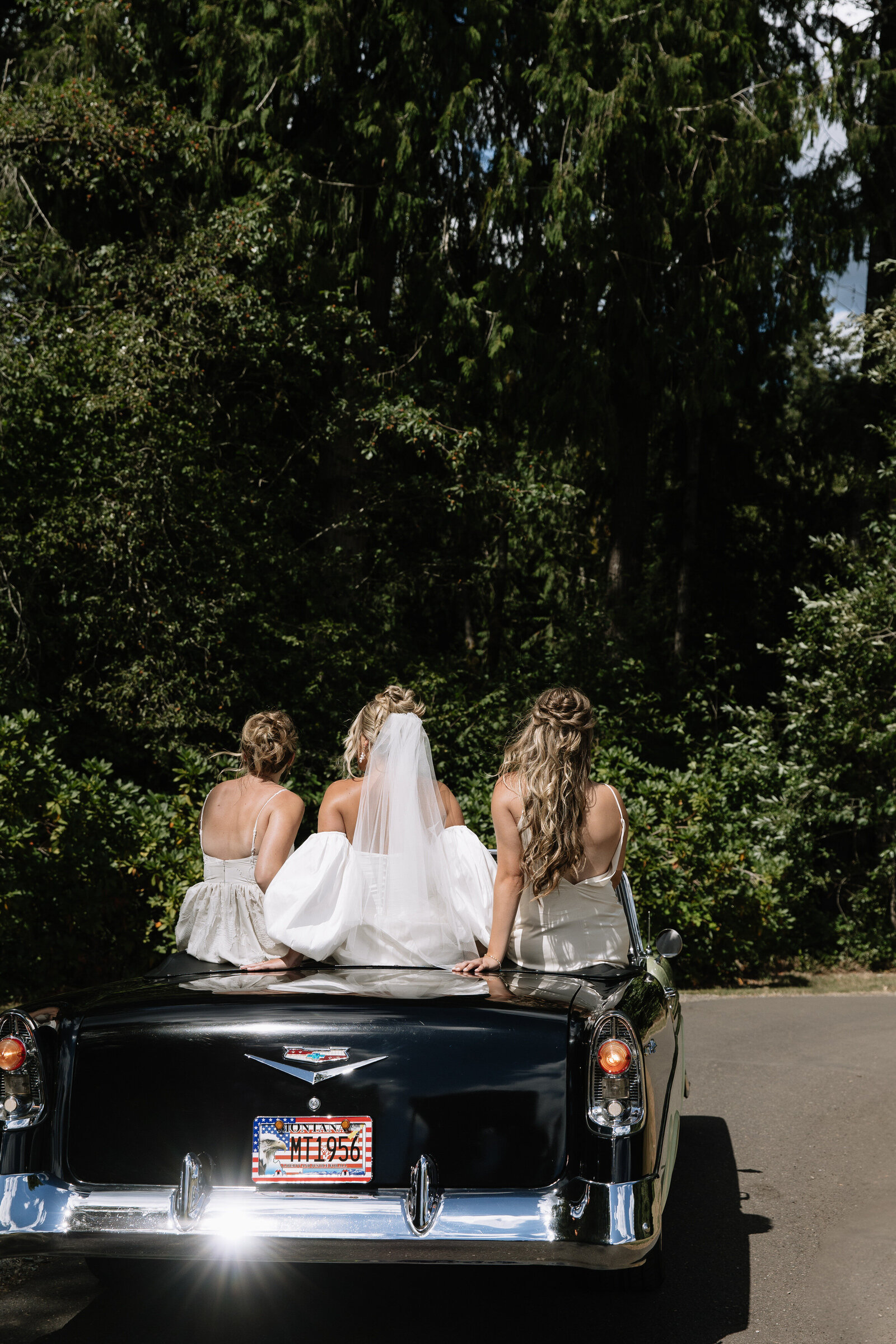 Bride and bridesmaids sit atop convertible classic car on the way to outdoor Washington wedding ceremony