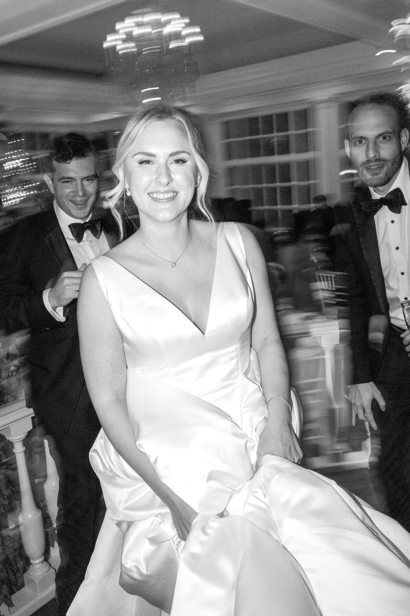 Black and white photo of a smiling bride in a wedding dress, walking inside a venue. Two men in tuxedos, one on each side, follow her. The setting is elegantly lit with chandeliers and large windows in the background.