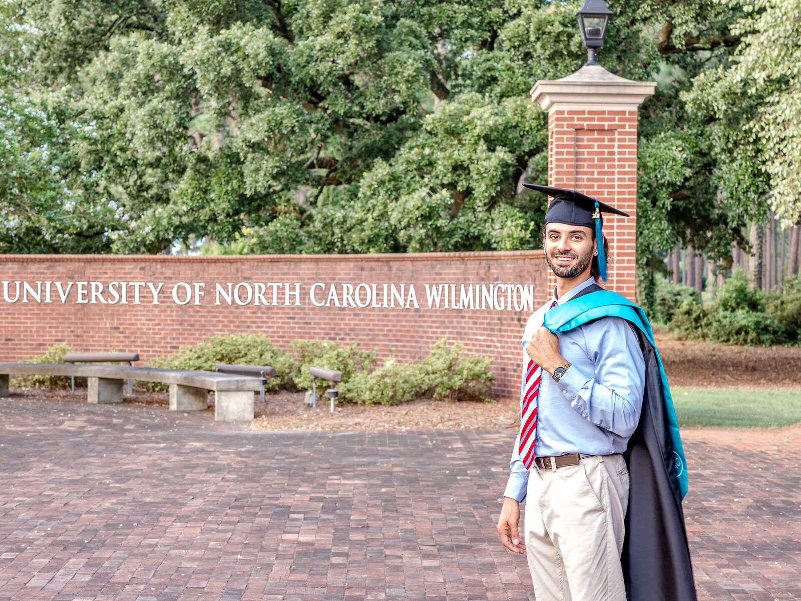 A proud senior graduate in cap and gown posing in front of the University of North Carolina Wilmington sign. This professional image captures the achievement of graduation, ideal for those seeking senior photography packages in Wilmington, NC.