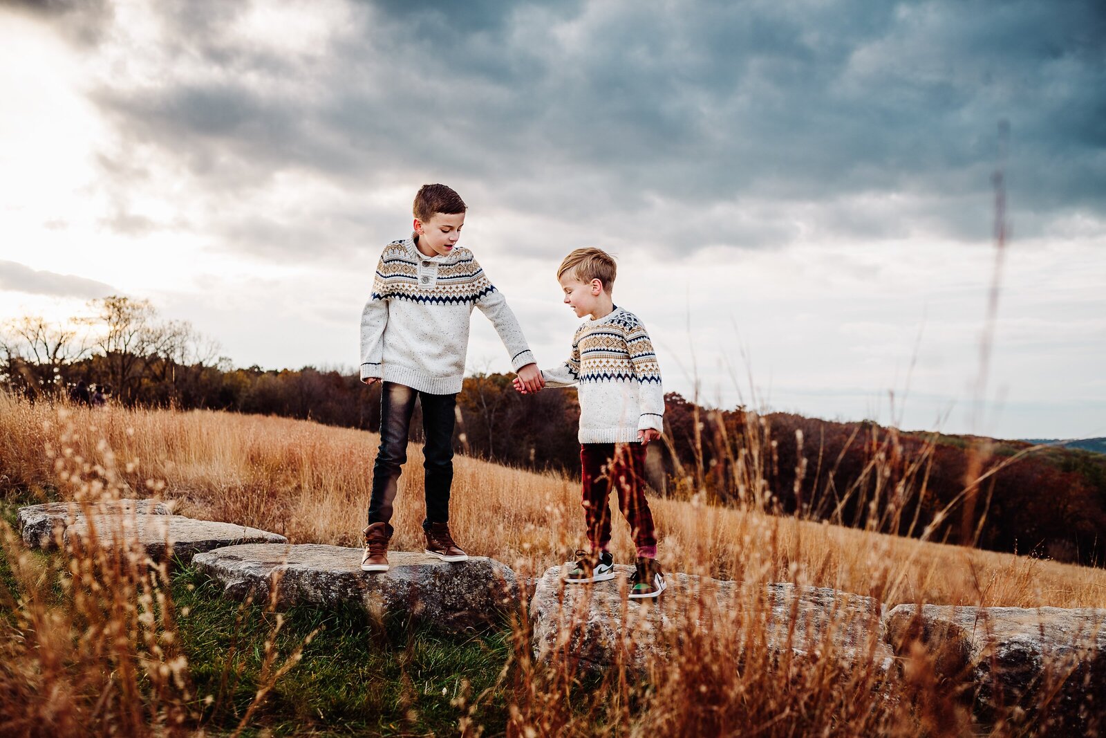 Two brothers in patterned sweaters, holding hands and standing on rocks with stormy skies behind them and golden wheat around them.