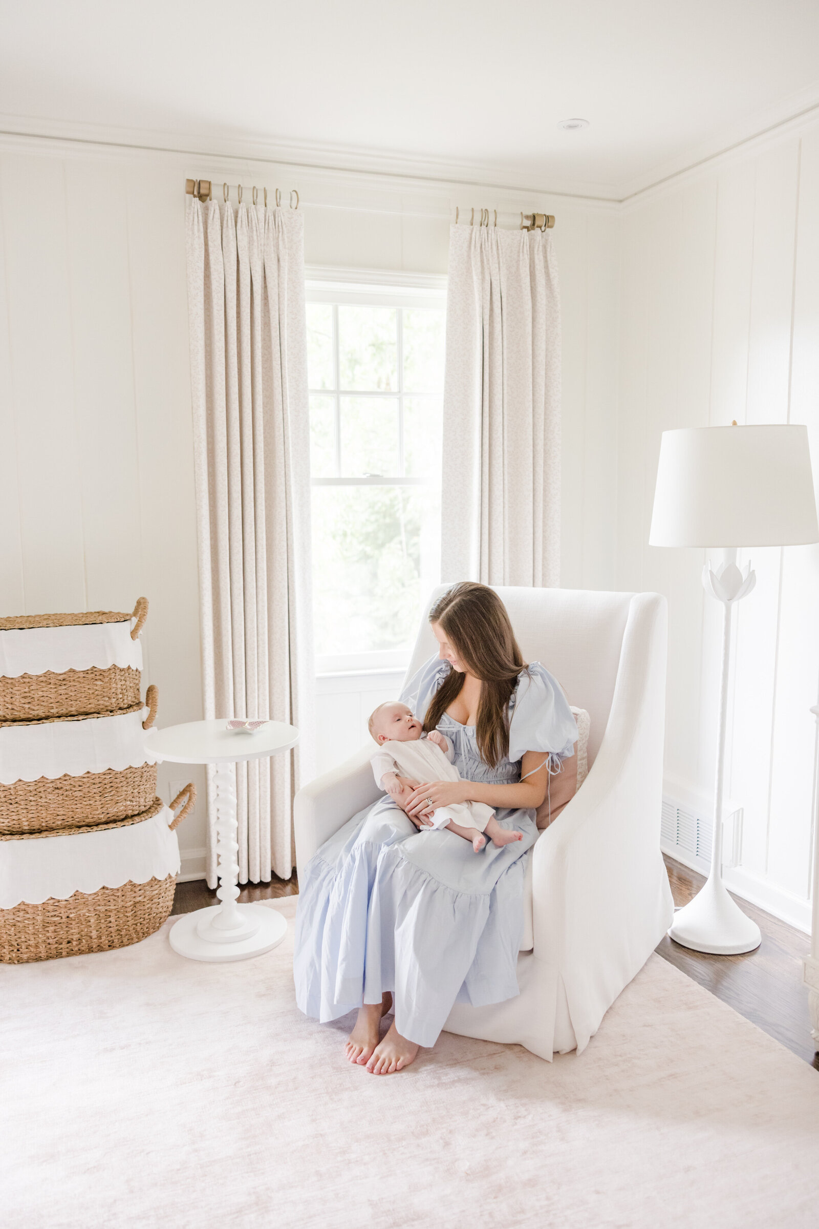 Mother seating in a white glider smiling down at newborn in her arms.