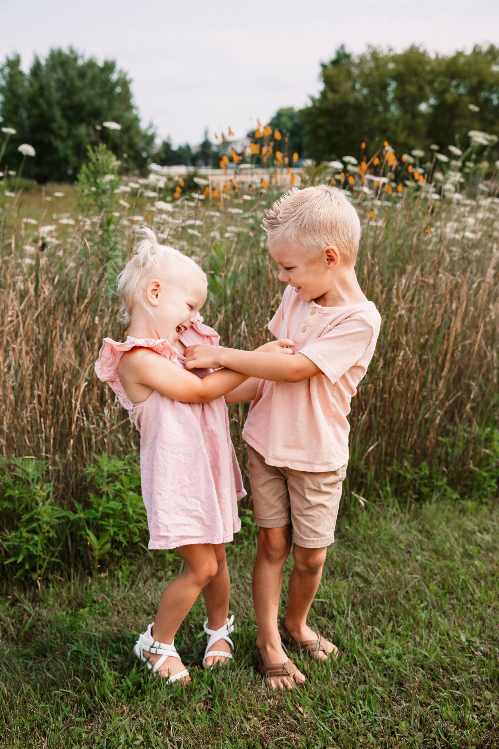 Happy toddler brother and sister in pink tickle each other while standing in a path with wildflowers