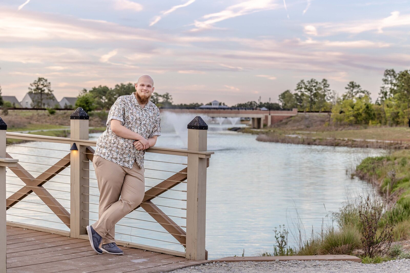 A Cape Fear Community College senior leaning on a wooden bridge by a scenic lake at sunset in North Carolina. This relaxed portrait captures the essence of professional senior photography, ideal for those seeking natural and serene photos in Wilmington.